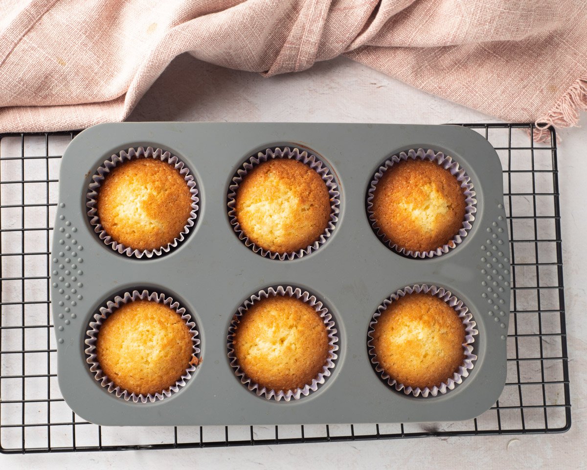 A person is spooning batter into cupcake liners in a gray silicone baking tray, suggesting the preparation of gluten free vanilla cupcakes for Easter or spring-themed celebrations.
