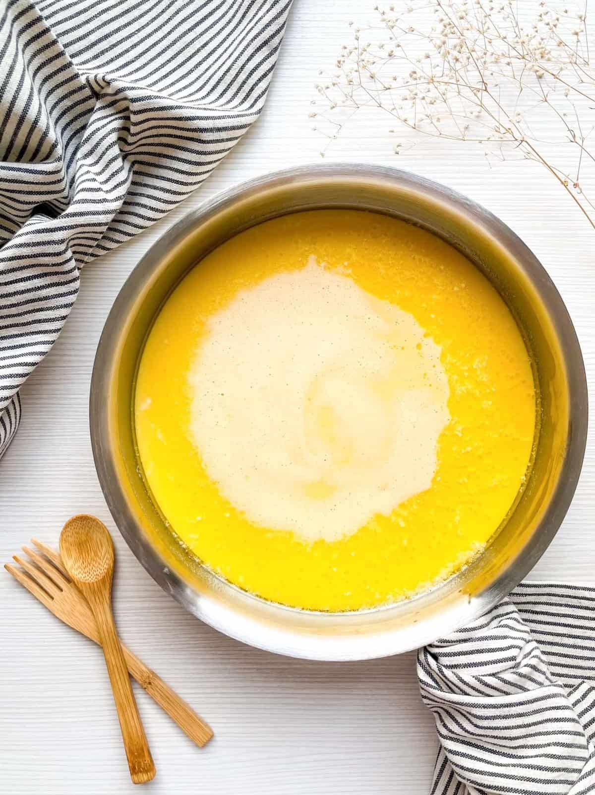 A pan filled with a yellow batter for a Dutch baby pancake recipe is placed on a white surface alongside bamboo utensils and a striped cloth.
