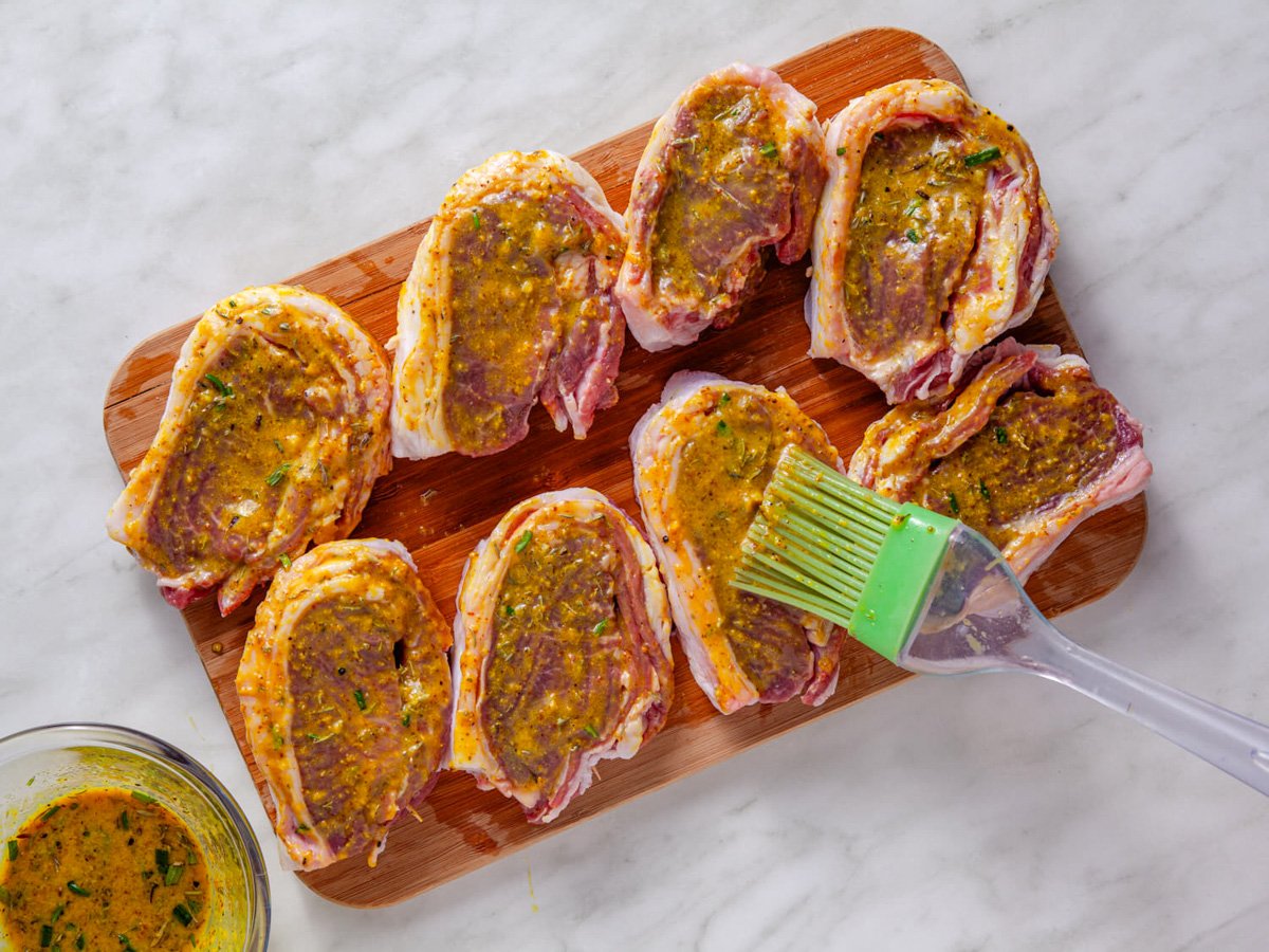 A wooden cutting board holds several lamb chops coated with a yellow marinade, along with a small bowl of the marinade and a basting brush.
