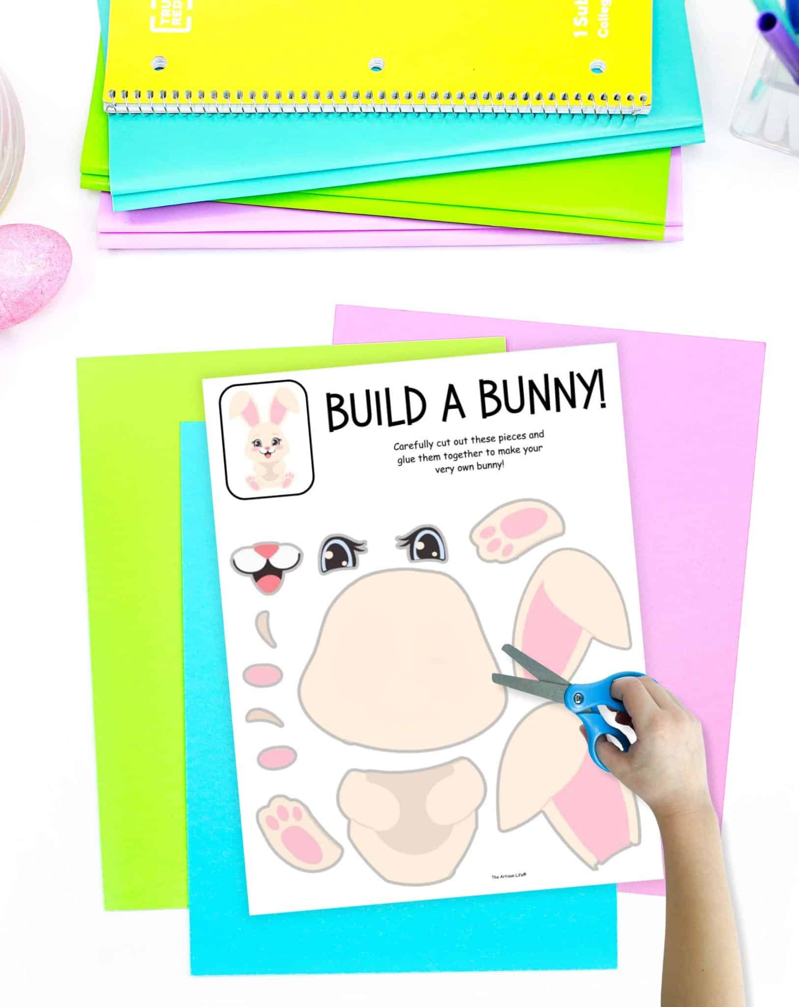 A child's hand holding scissors cuts out pieces from a "Build a Bunny" printable worksheet, surrounded by colorful sheets of paper, demonstrating a spring craft activity suitable for preschool and kindergarten students.
