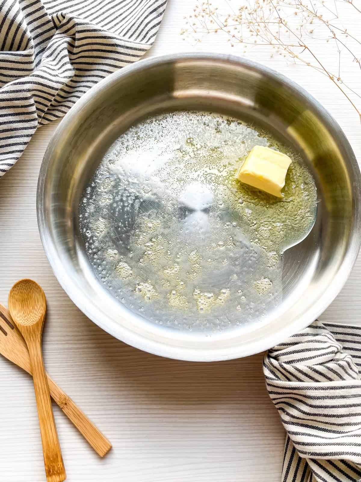 A stainless steel pan contains melted butter and bubbling liquid, surrounded by wooden utensils and a striped kitchen towel, suggesting preparations for a Dutch baby pancake recipe.
