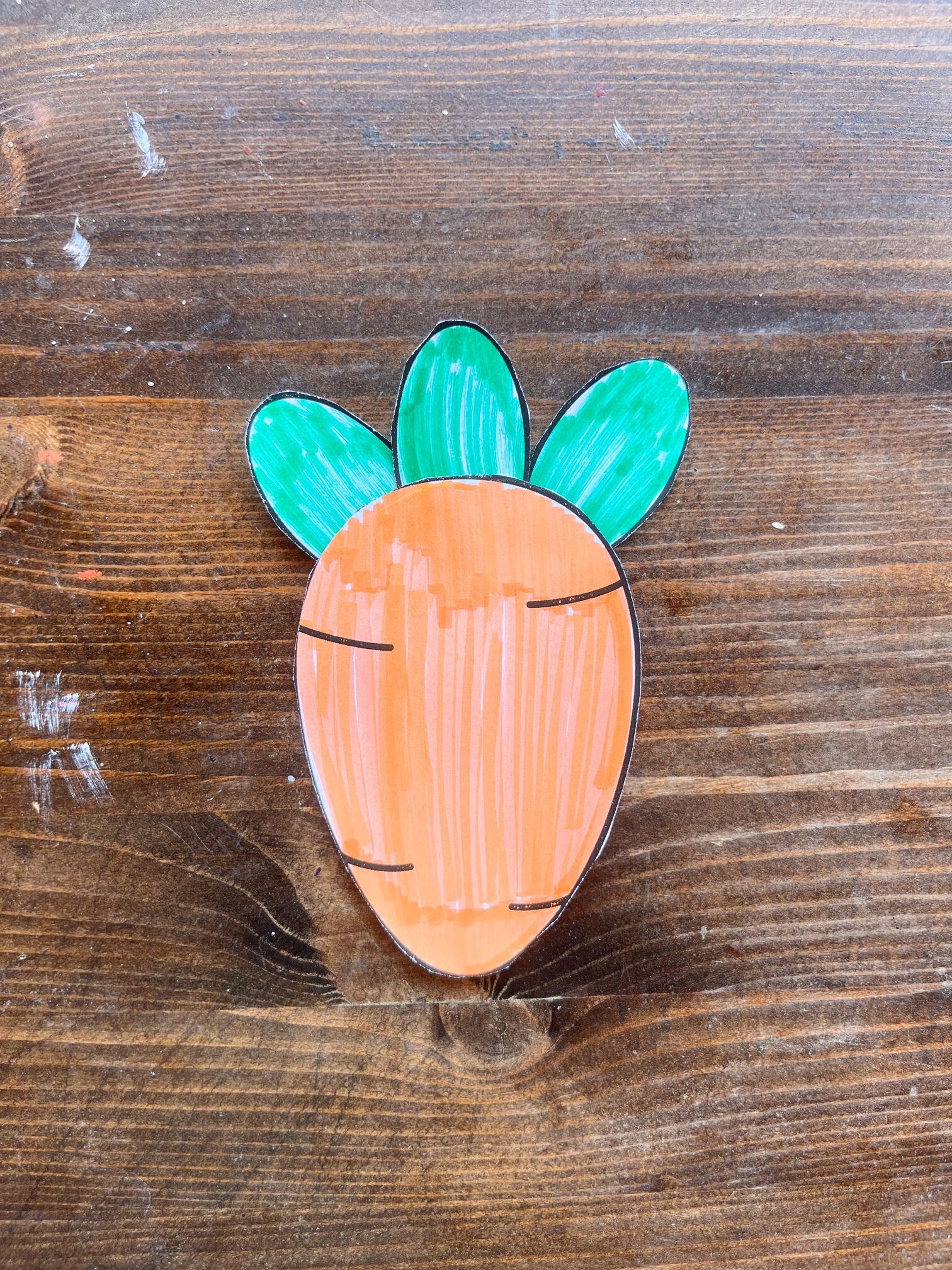 A hand-drawn paper carrot with green leaves is placed on a wooden surface, serving as a DIY craft idea for Easter activities, suitable for toddlers and preschoolers.
