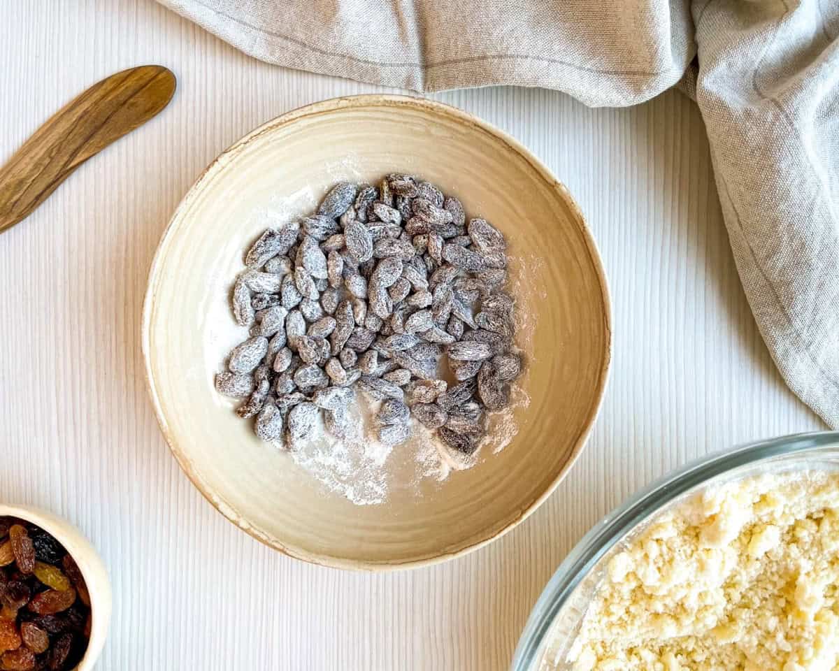 A bowl of mixed raisins coated in flour sits next to a glass bowl of flour, with a wooden spoon and cloth in the background, suggesting preparation for an easy buttermilk scone or Irish soda bread recipe.
