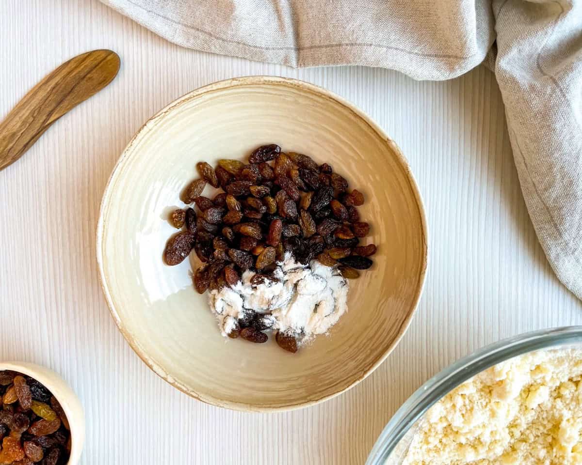 A bowl of floured raisins sits alongside a bowl of mixed ingredients, suggesting preparation for making buttermilk scones or Irish soda bread scones.
