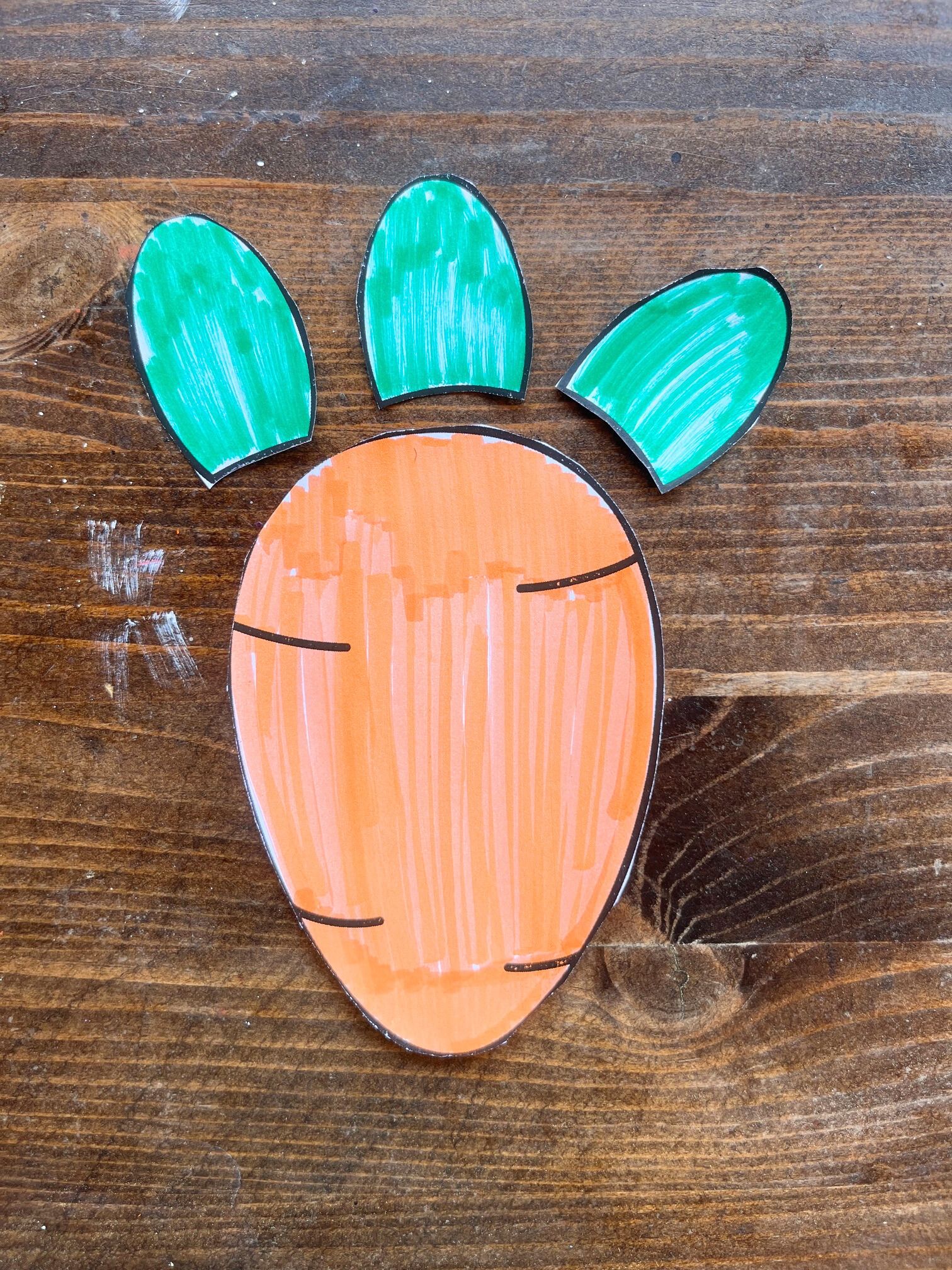 A colorful paper carrot cutout with green tops, resting on a wooden surface, suitable for DIY Easter crafts, carrot crafts, and spring activities for kids.
