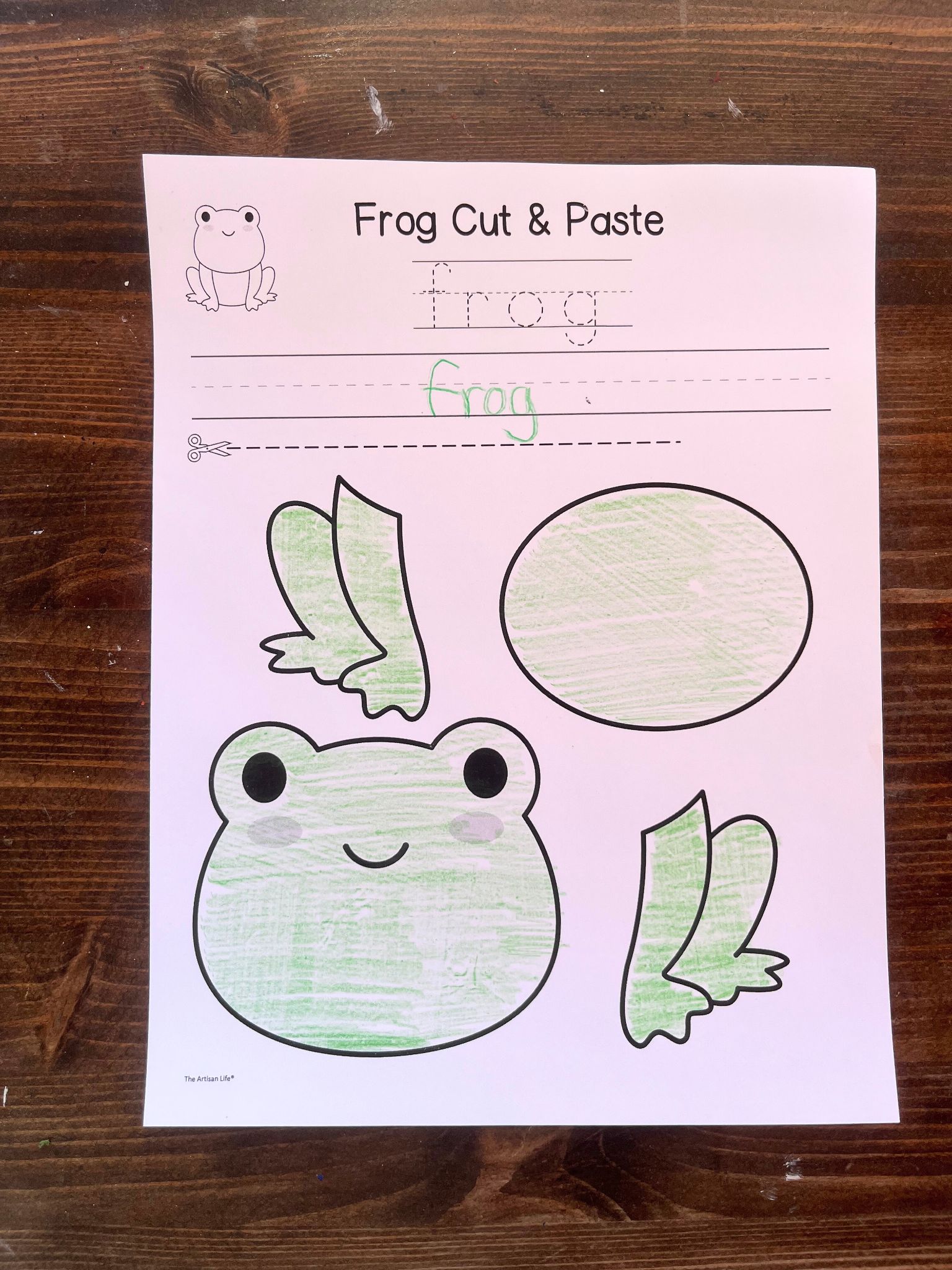 A printed activity sheet for kids titled "Frog Cut & Paste" features a frog template with sections for cutting out a frog's body and legs, alongside a space for practicing the word "frog."
