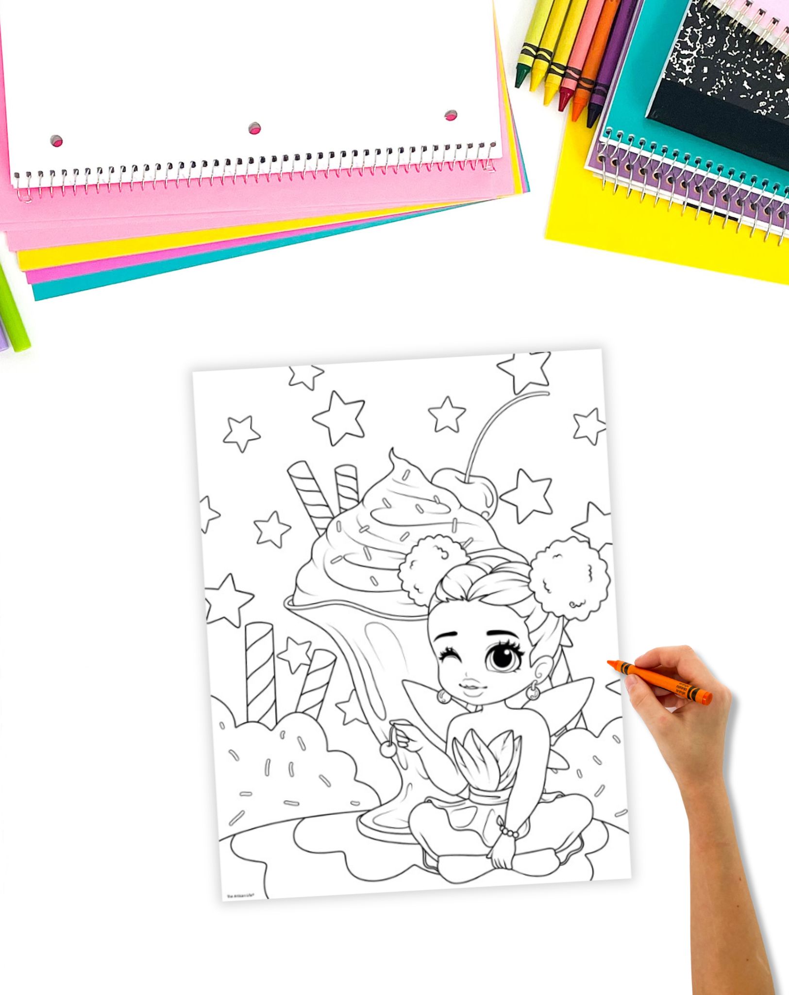 A child is coloring a fairy-themed page featuring a magical fairy sitting beside an ice cream sundae, surrounded by stars and colorful dessert elements, with various coloring supplies nearby.
