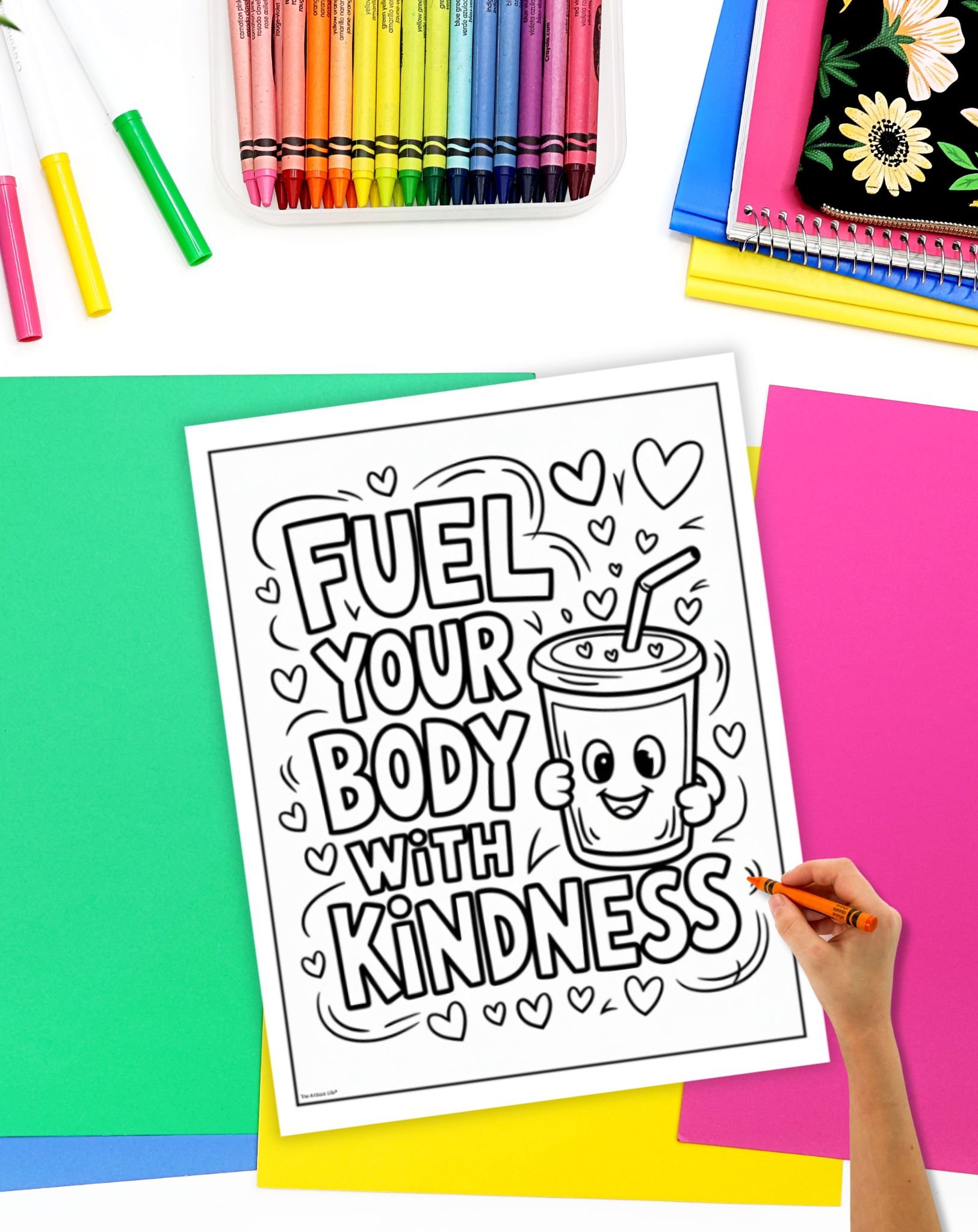 A hand is holding a crayon, coloring a black-and-white printable page featuring the quote "FUEL YOUR BODY WITH KINDNESS," surrounded by colorful sheets of paper and a container of crayons in the background.
