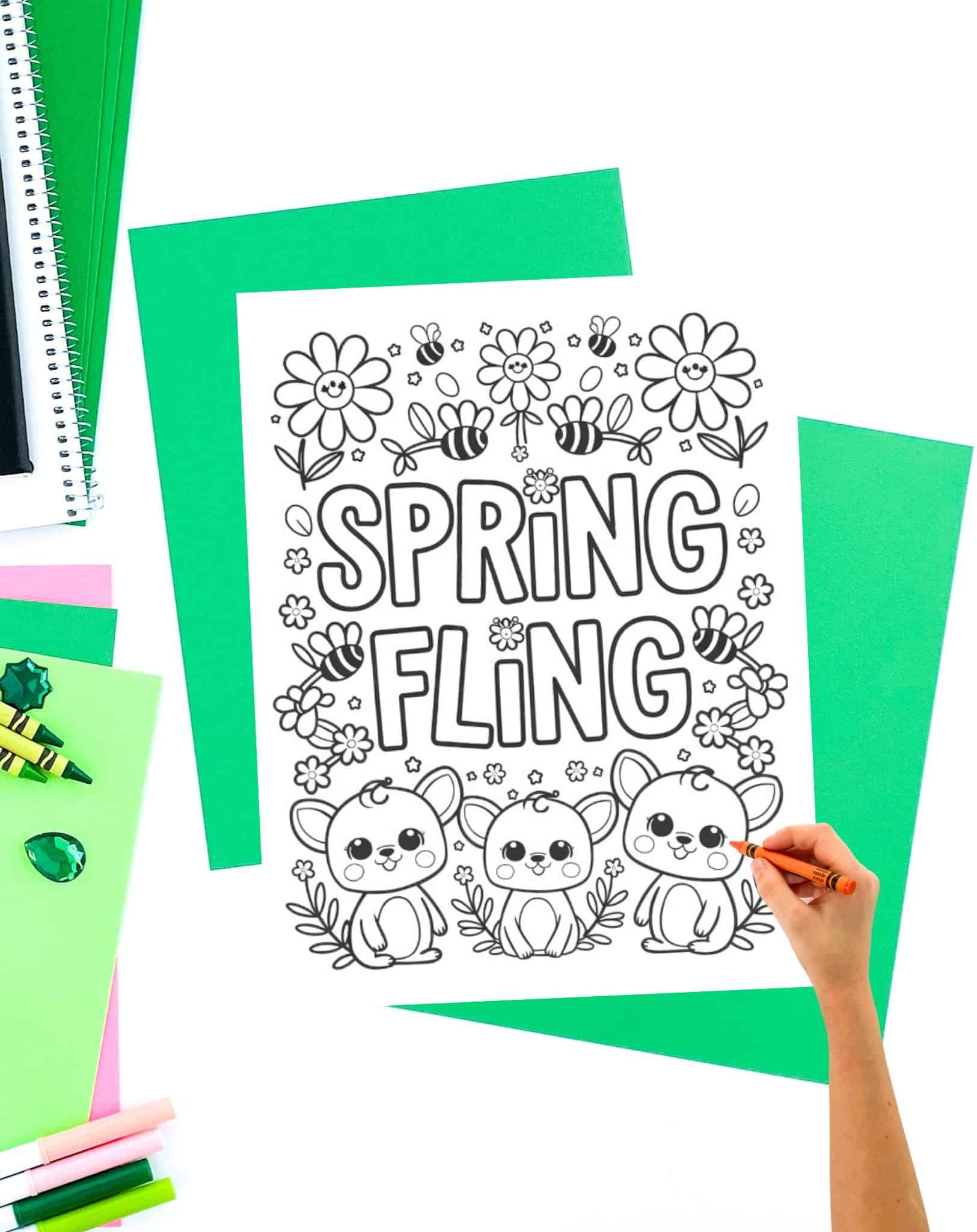 A hand colors a printable Easter-themed coloring page featuring the text "SPRING FLING" along with cute bunny illustrations and flowers, surrounded by various colored papers and crayons.
