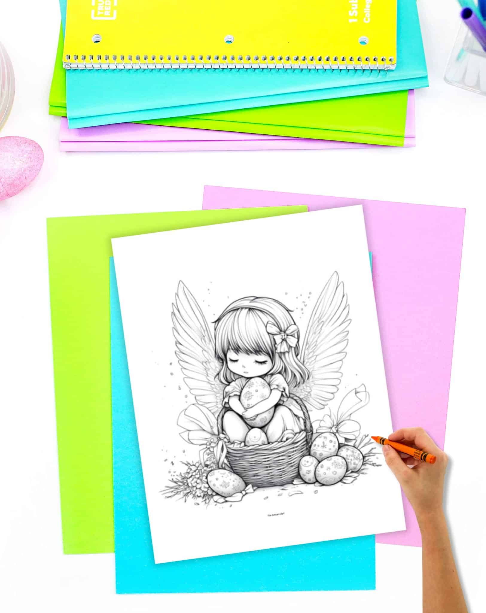 A hand holds an orange crayon over a black-and-white Easter coloring page depicting a girl with wings sitting in a basket surrounded by decorated eggs, set against a backdrop of colorful sheets of paper and a small decorative egg.
