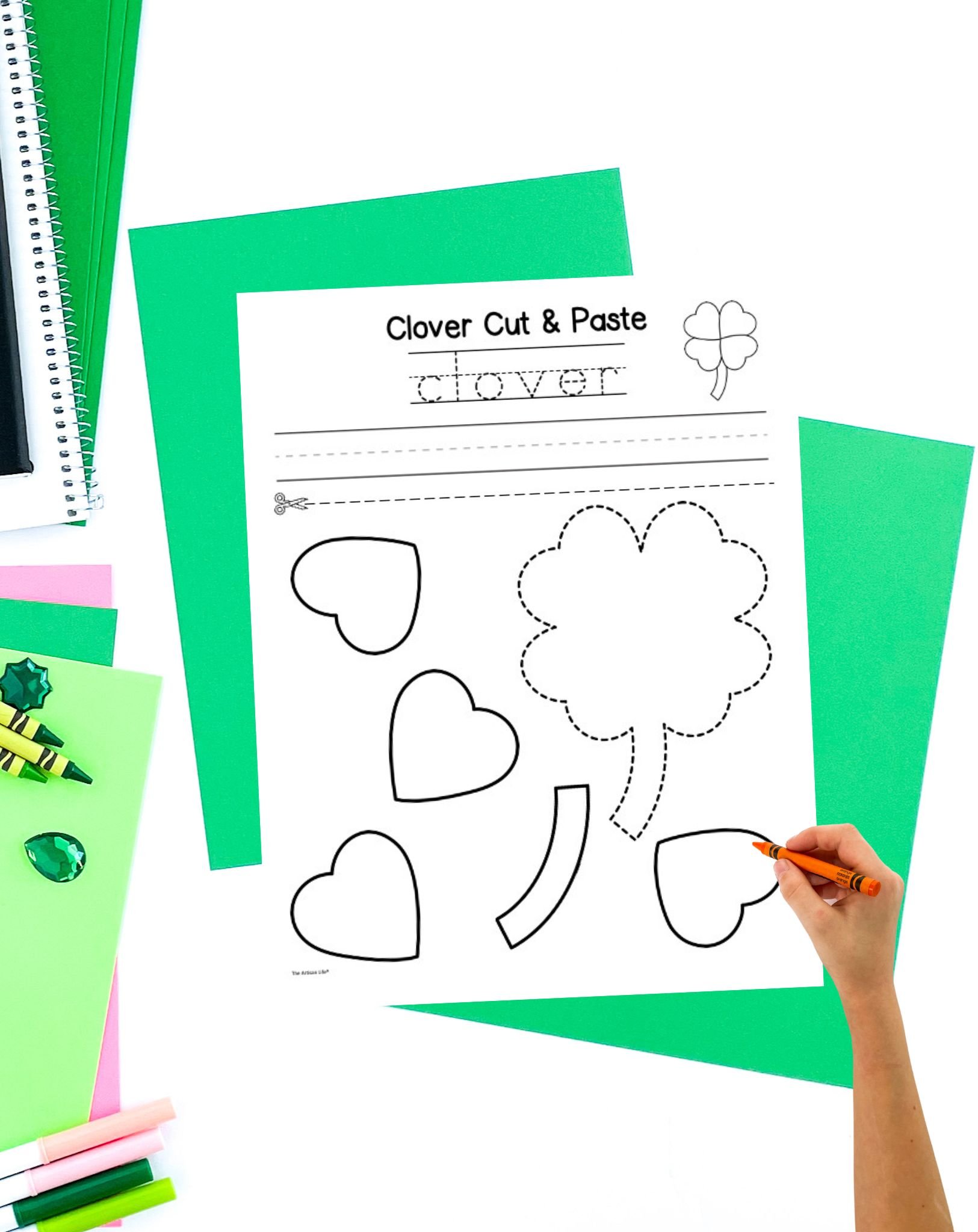 A hand is coloring a clover cut-and-paste activity sheet, surrounded by green and pink construction paper, markers, crayons, and decorative gems, highlighting a four leaf clover template ideal for St. Patrick’s Day crafts for kids and toddlers.
