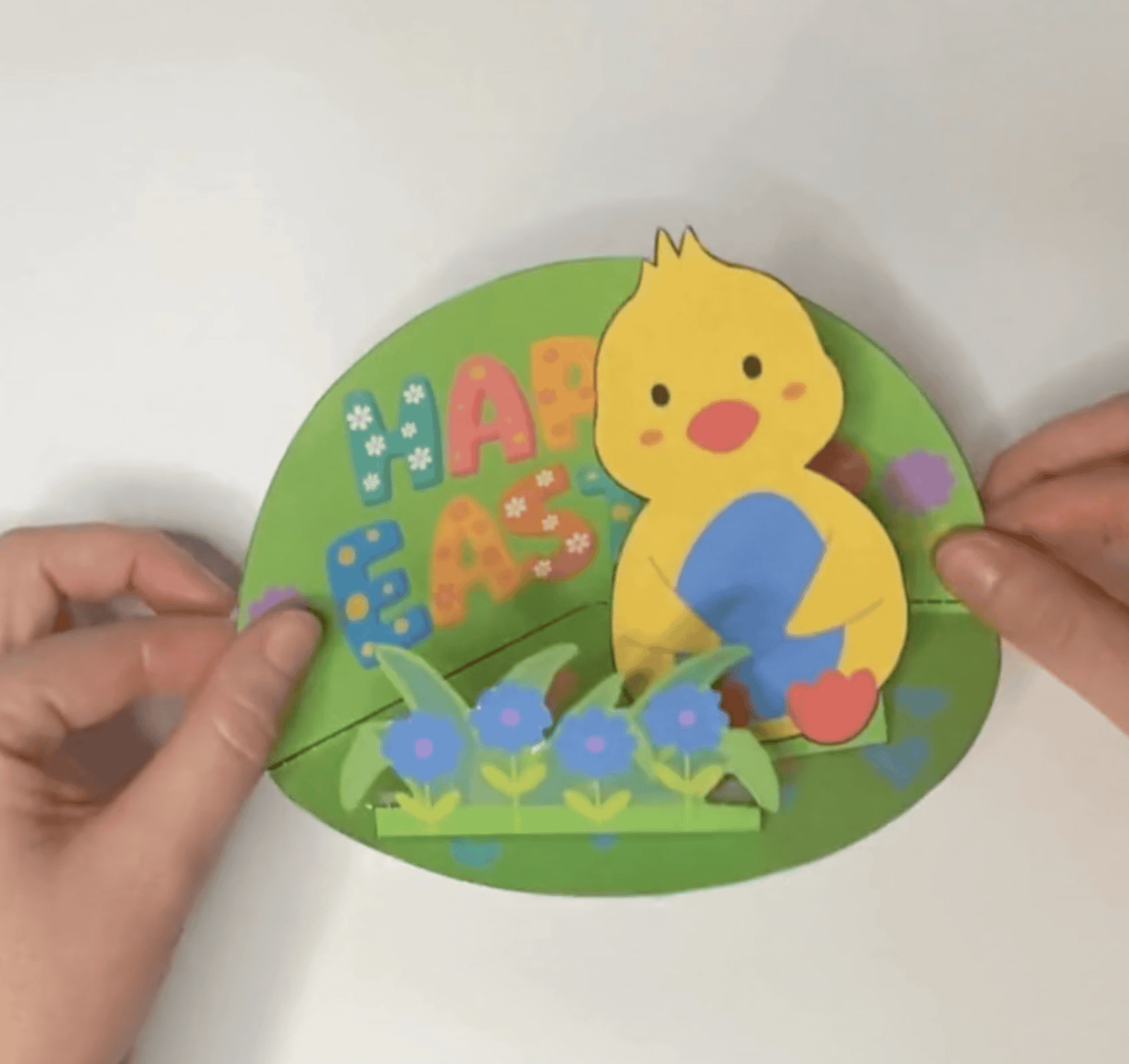 A person is holding a colorful, handmade Easter card featuring a chick and flowers, designed as a DIY project for kids, suitable for preschool and kindergarten crafts.
