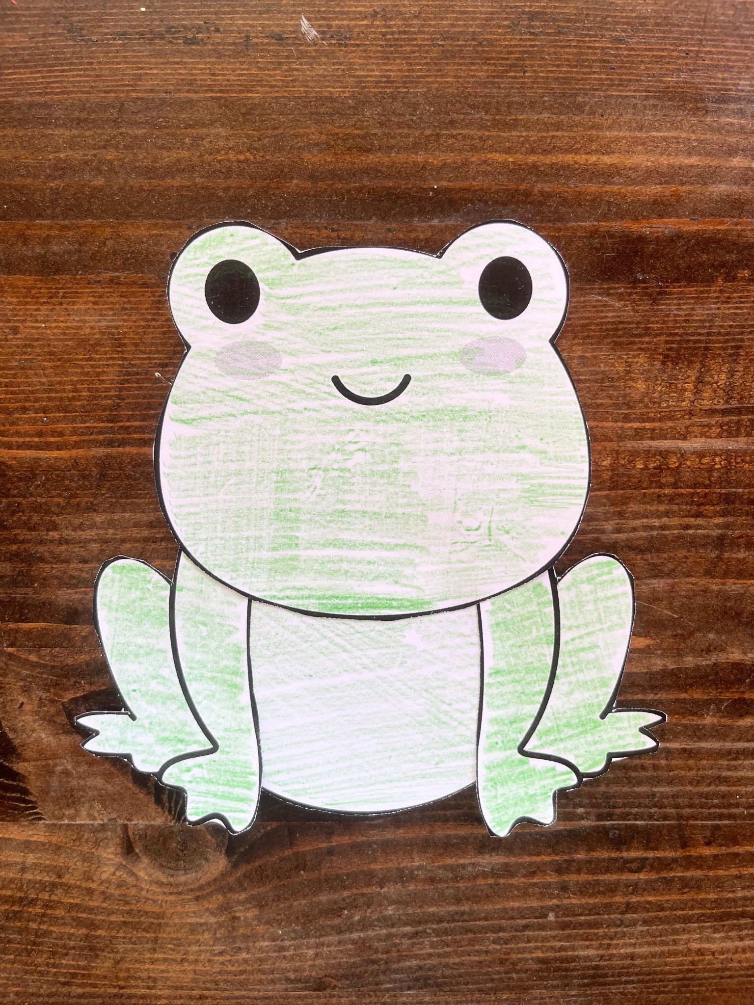 A cutout of a cartoon frog, suitable for preschool activities, is displayed on a wooden surface, ideal for various frog-themed crafts for kids.

