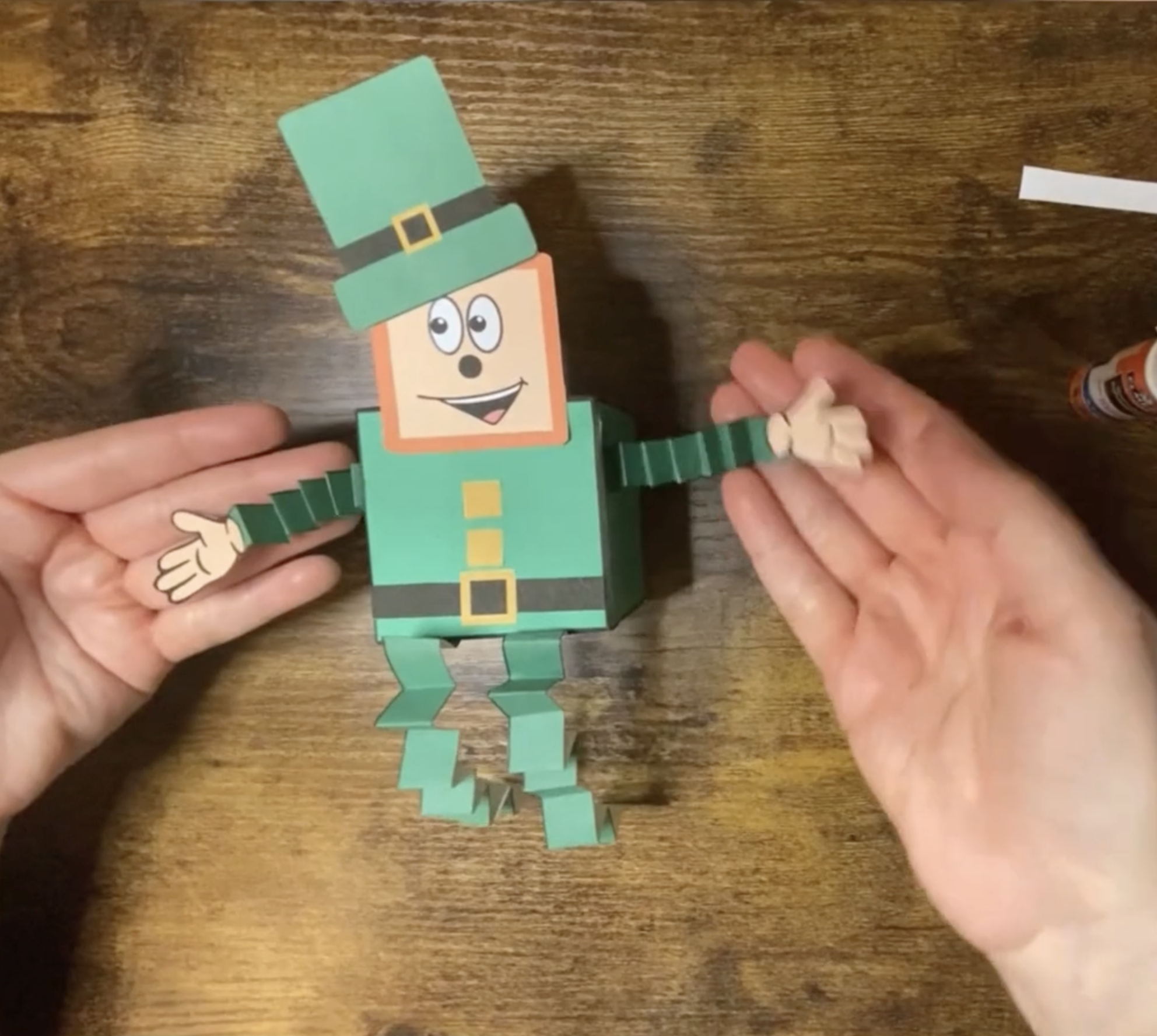 A person is assembling a leprechaun craft using green and brown paper pieces and glue, suitable for St. Patrick’s Day activities for children.
