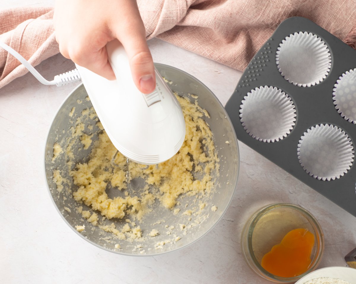 A person using a hand mixer is preparing a gluten-free vanilla cupcake batter, holding a bowl with an egg yolk above a mixing bowl that contains a creamy mixture, with cupcake liners and baking ingredients visible in the background.
