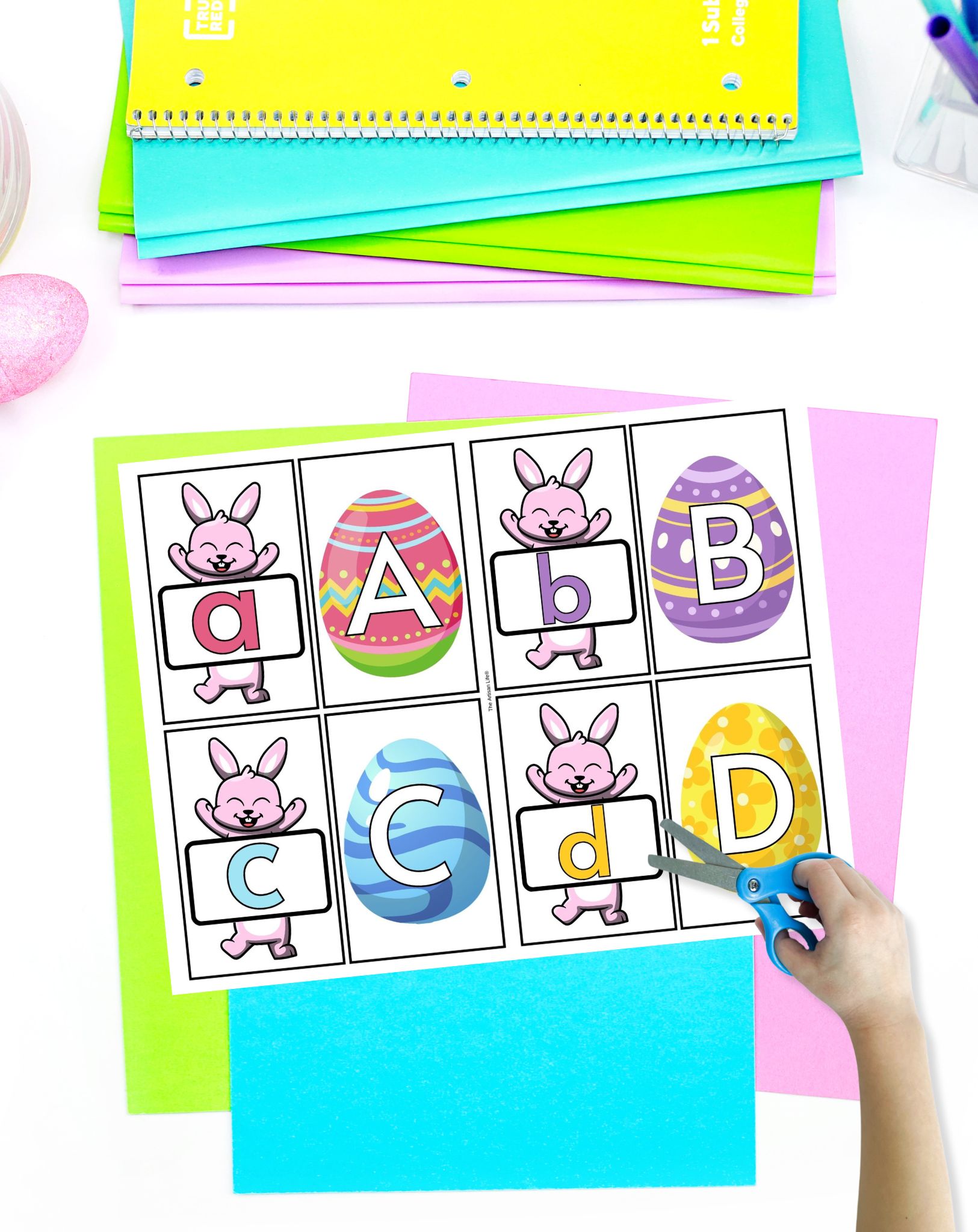 A child's hand holds scissors as they cut out Easter-themed letter cards featuring bunnies and decorated eggs, surrounded by colorful paper, illustrating Easter alphabet activities for preschool.
