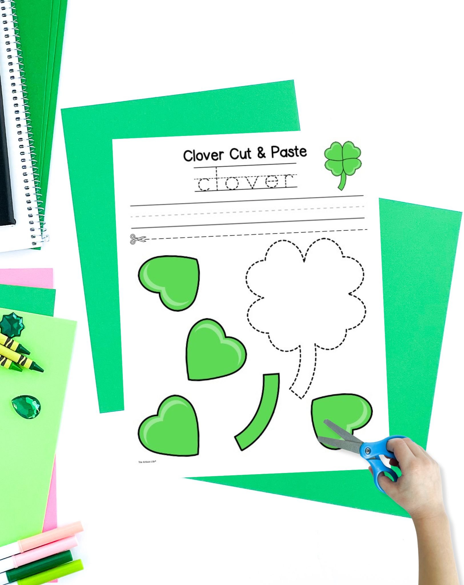 A hand with scissors is cutting out shapes from a "Clover Cut & Paste" worksheet featuring a four leaf clover template, surrounded by colorful paper and craft supplies for a St. Patrick's Day craft activity.
