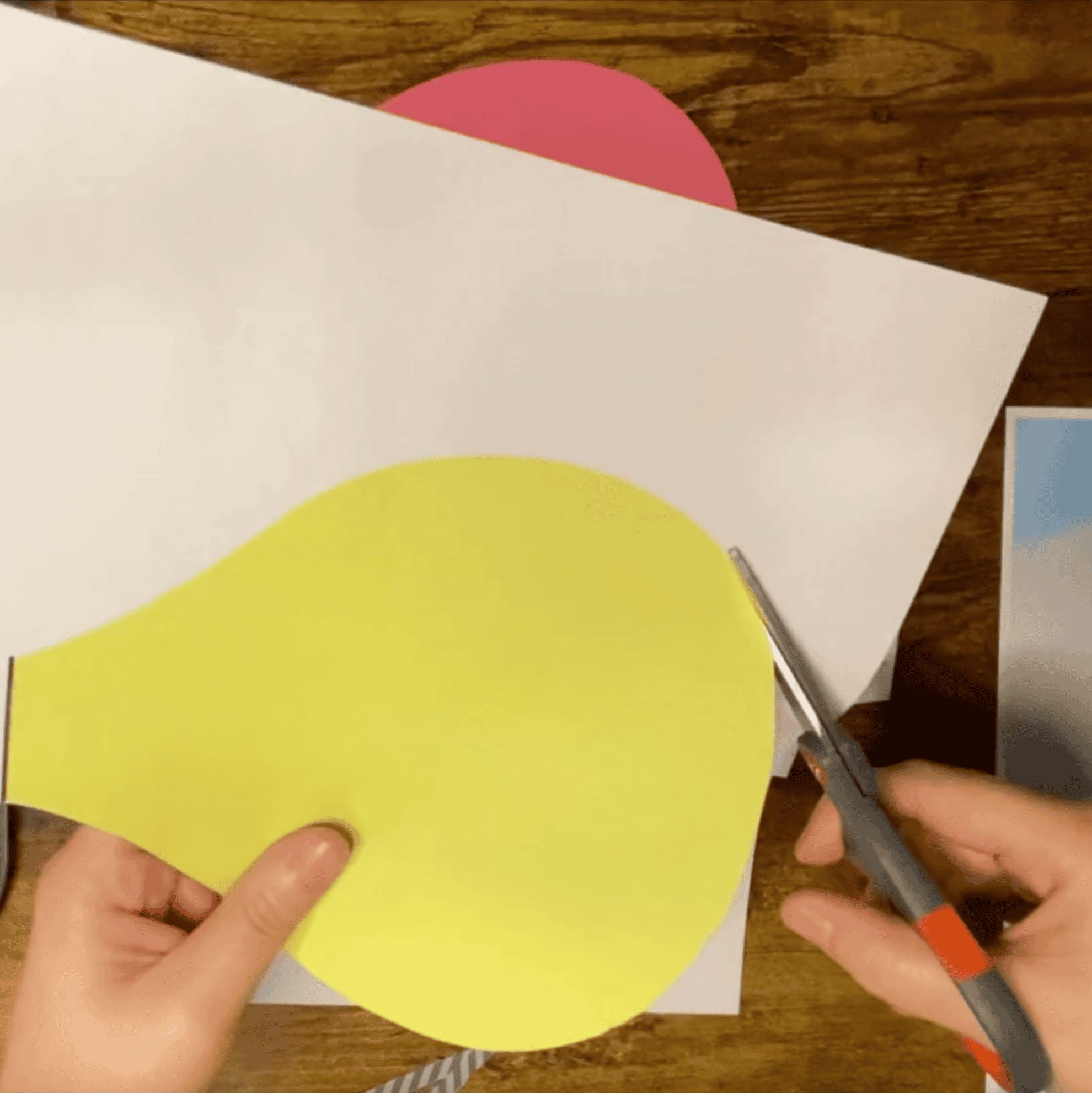 A person is cutting a yellow paper hot air balloon shape using scissors, with additional colored paper and templates visible in the background, suggesting a hot air balloon craft project for preschoolers.
