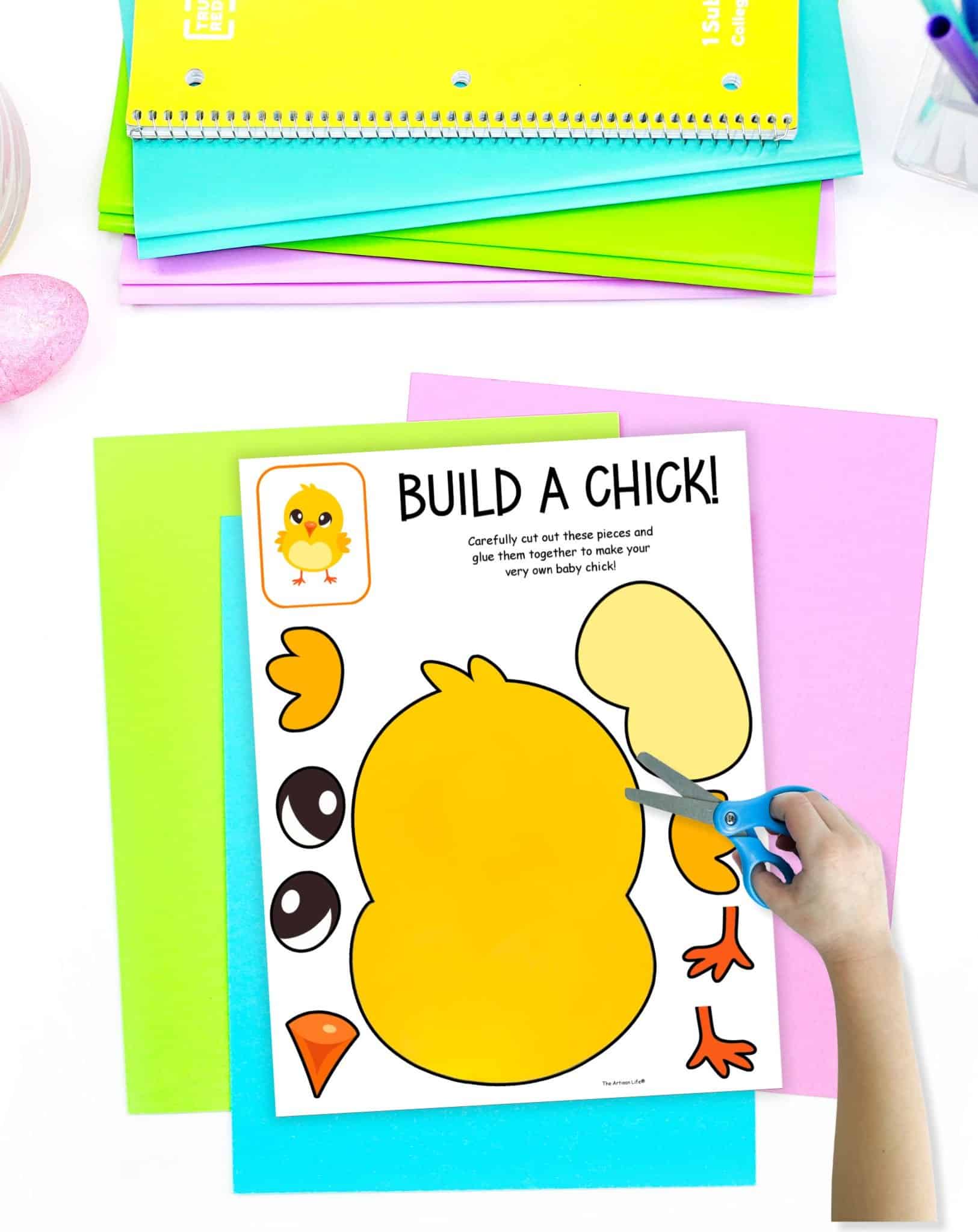 A child's hand holds scissors over a "Build a Chick" cut-and-paste worksheet featuring pieces to create a cartoon chick, placed on colorful sheets of paper, ideal for spring crafts and printable Easter activities for kids.
