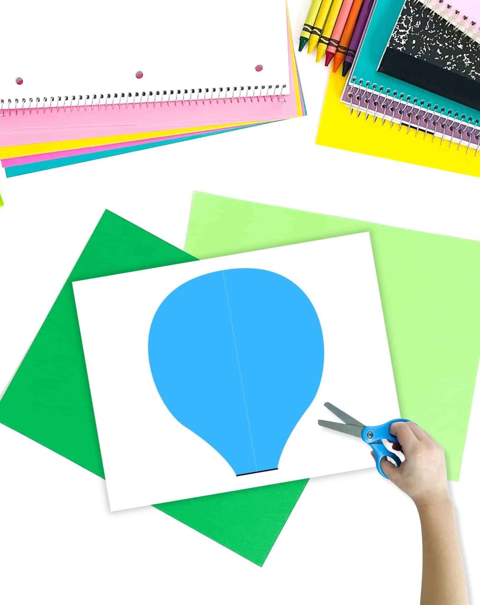 A hand holding scissors prepares to cut out a blue paper hot air balloon shape from a white sheet, surrounded by colorful paper and notebooks, illustrating a hot air balloon craft project suitable for preschool activities.
