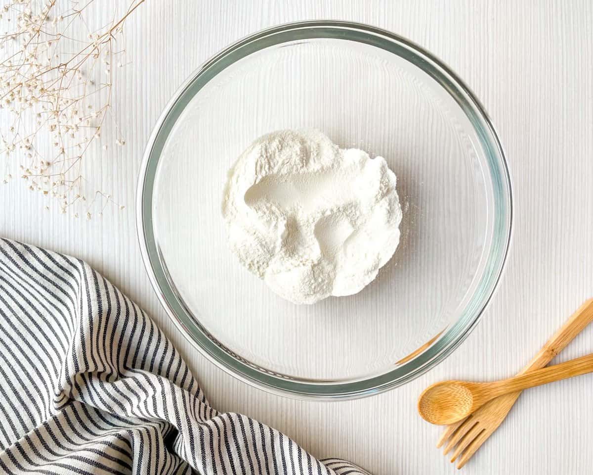 A clear glass bowl containing a mound of flour is placed on a white wooden surface, accompanied by a striped cloth, wooden utensils, and dried flowers, suggesting preparation for a Dutch baby pancake recipe.
