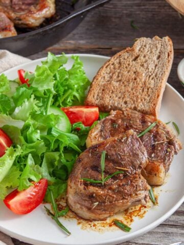 A plate featuring two seared lamb chops garnished with herbs, accompanied by a side salad of lettuce and tomatoes, a slice of bread, and a small dish of seasoning, with a cast iron skillet in the background.