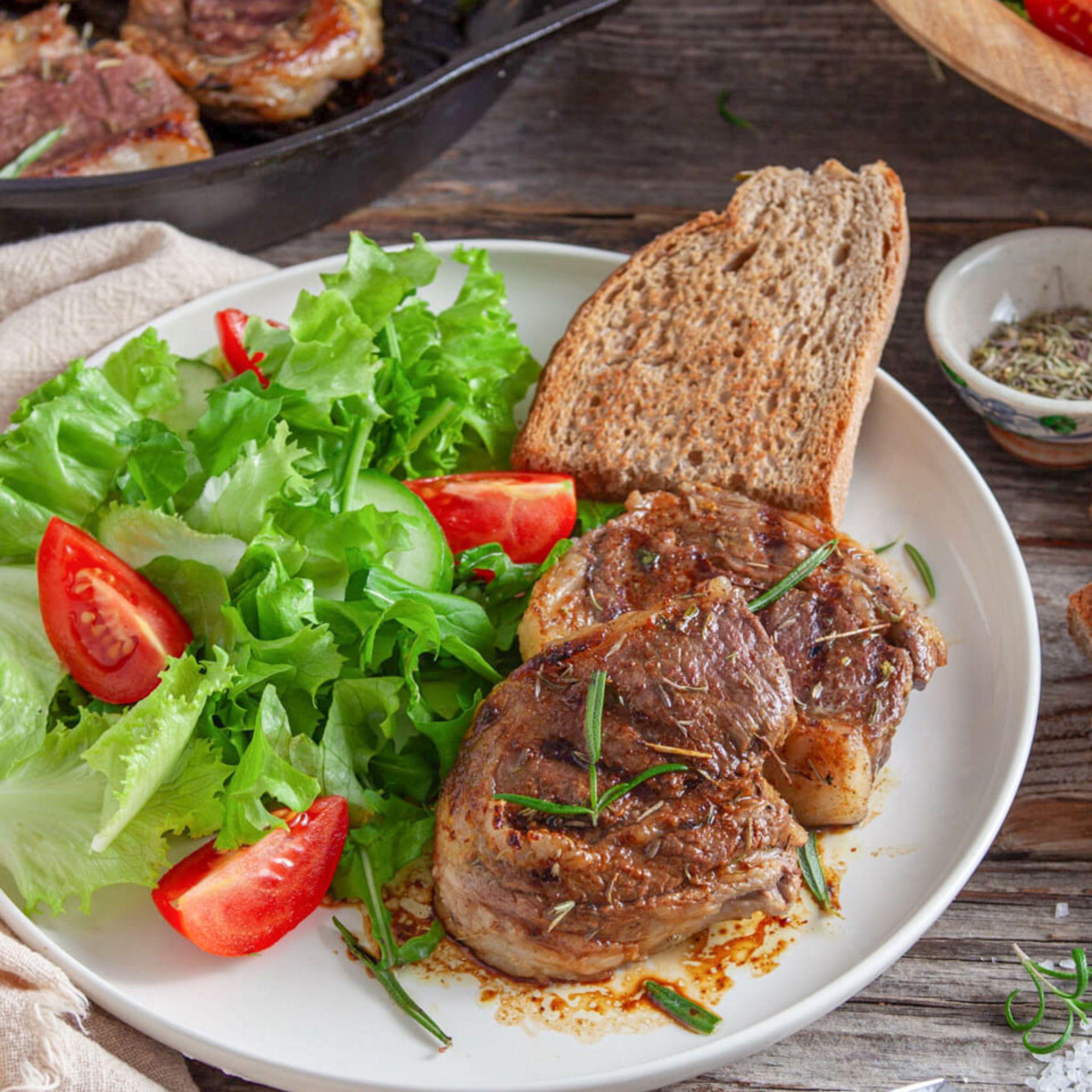 A plate featuring two seared lamb chops garnished with herbs, accompanied by a side salad of lettuce and tomatoes, a slice of bread, and a small dish of seasoning, with a cast iron skillet in the background.
