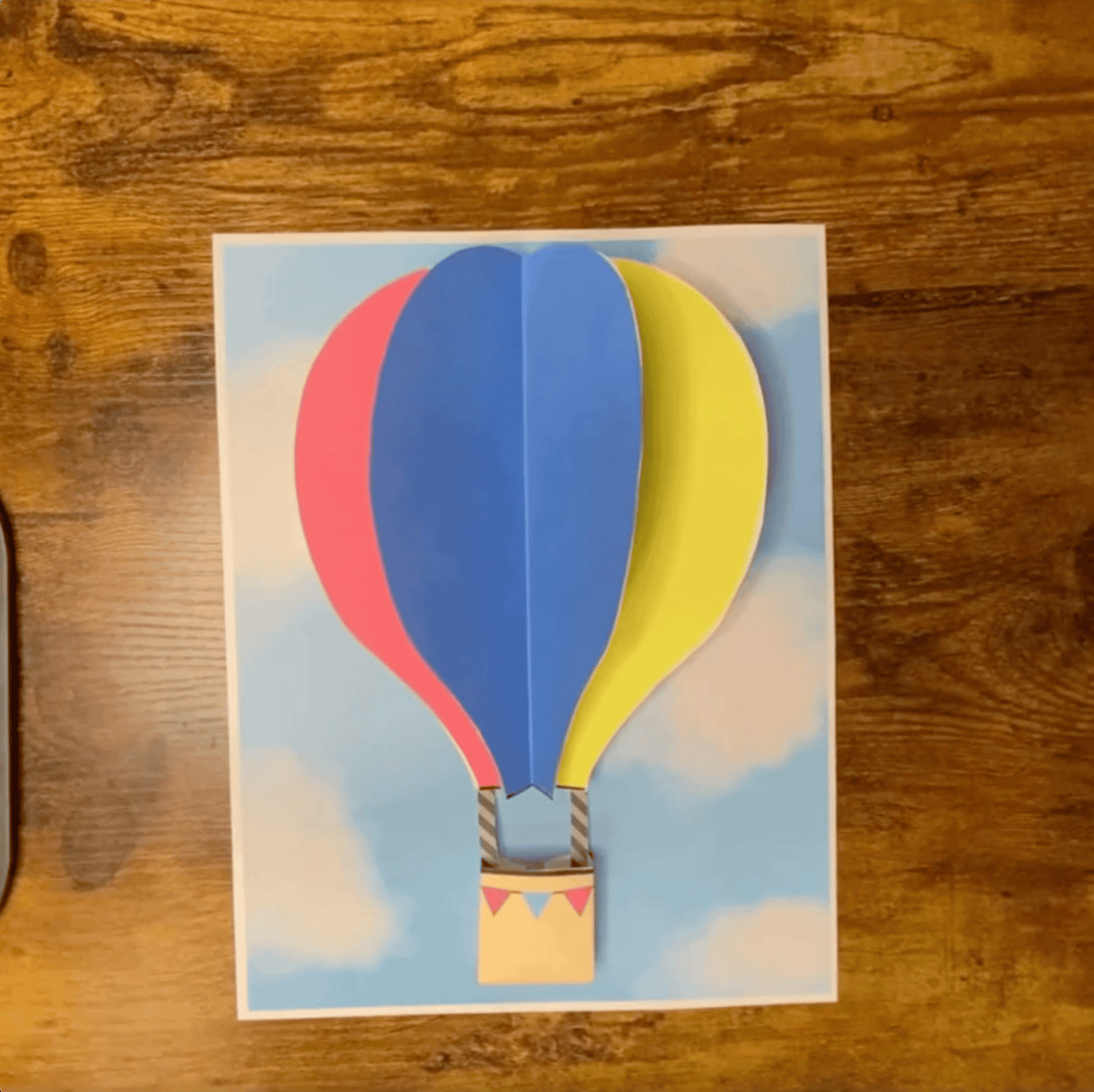 A paper hot air balloon craft with colorful balloon shapes is displayed against a sky background, illustrating a preschool art project.
