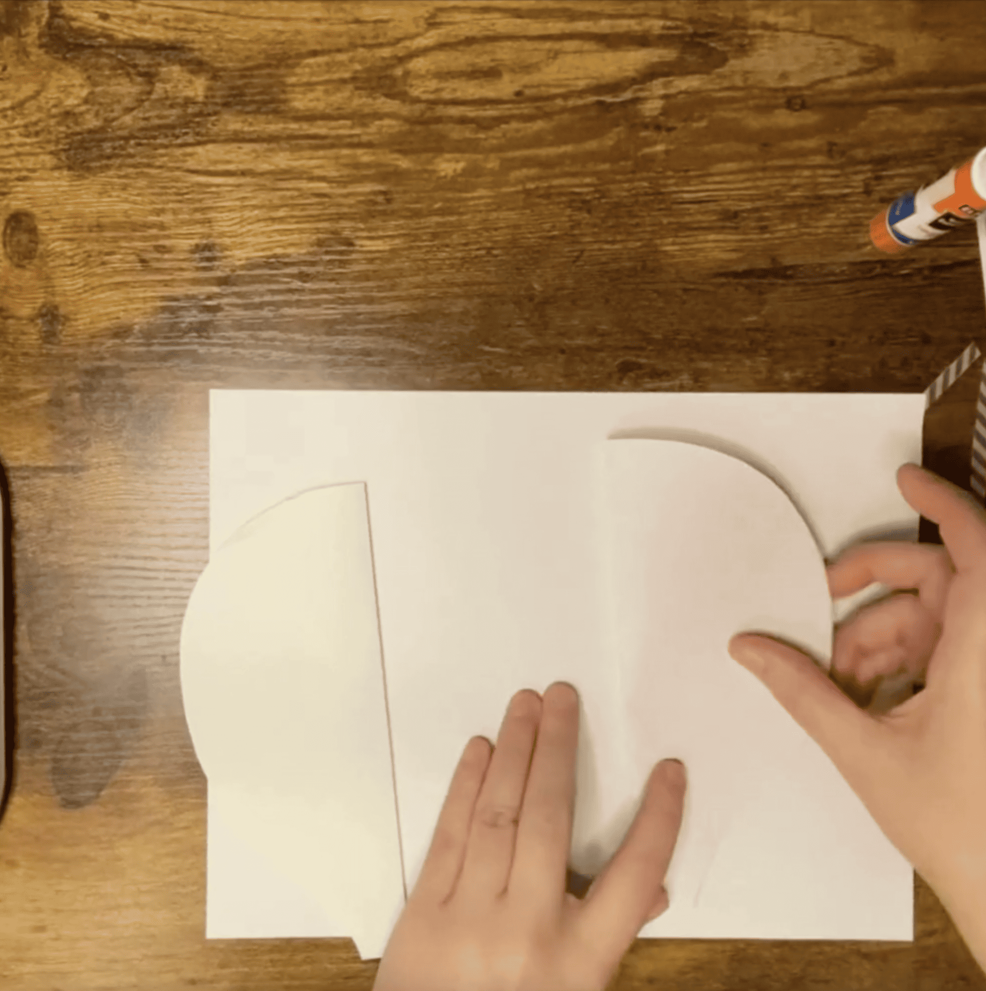 A person's hands are positioning cut paper shapes onto a blank sheet to create a hot air balloon craft for preschool art projects.
