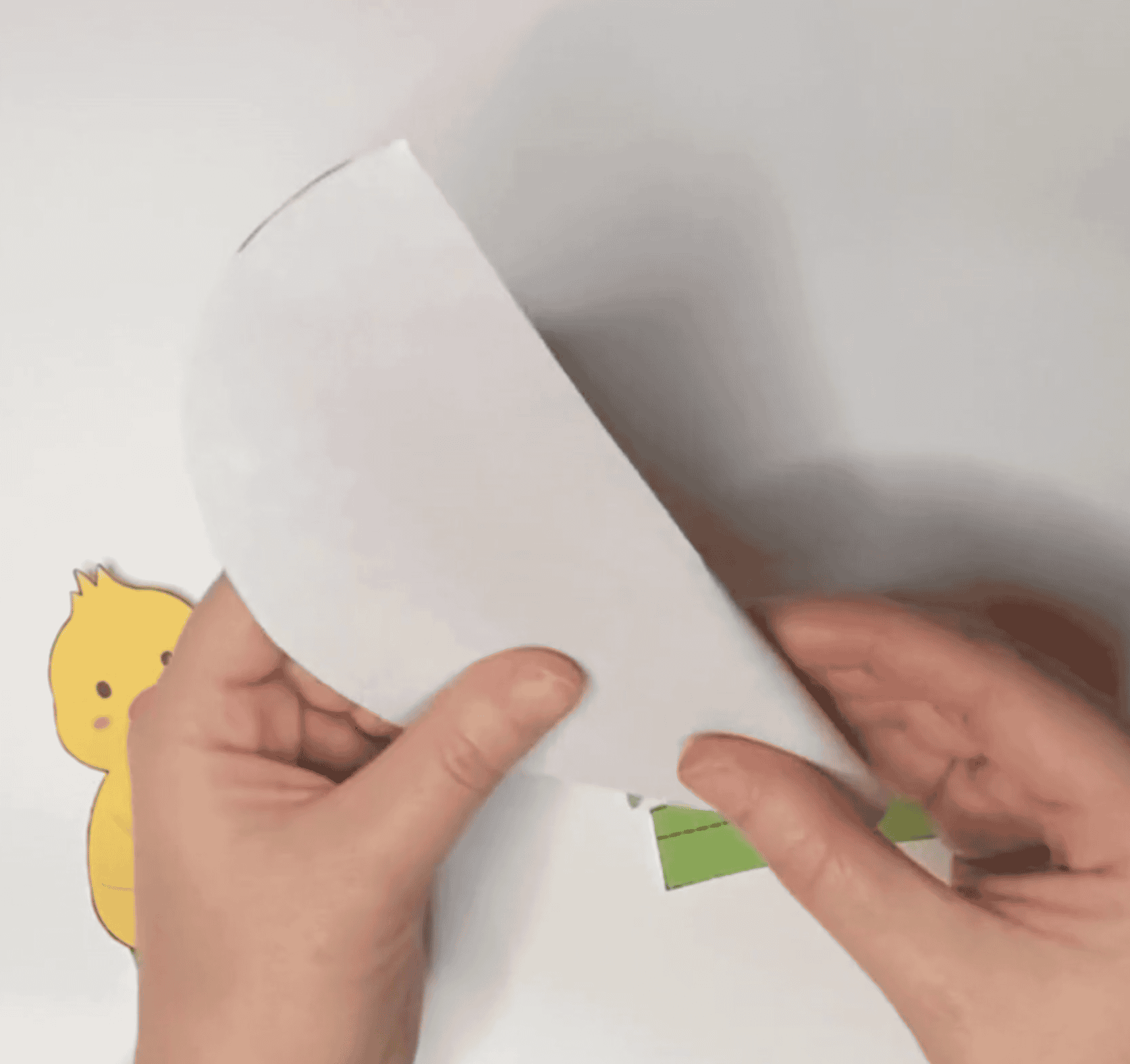 A person is folding a piece of paper to create a DIY Easter card, with a yellow chick and green paper pieces visible in the context of Easter-themed arts and crafts for kids.
