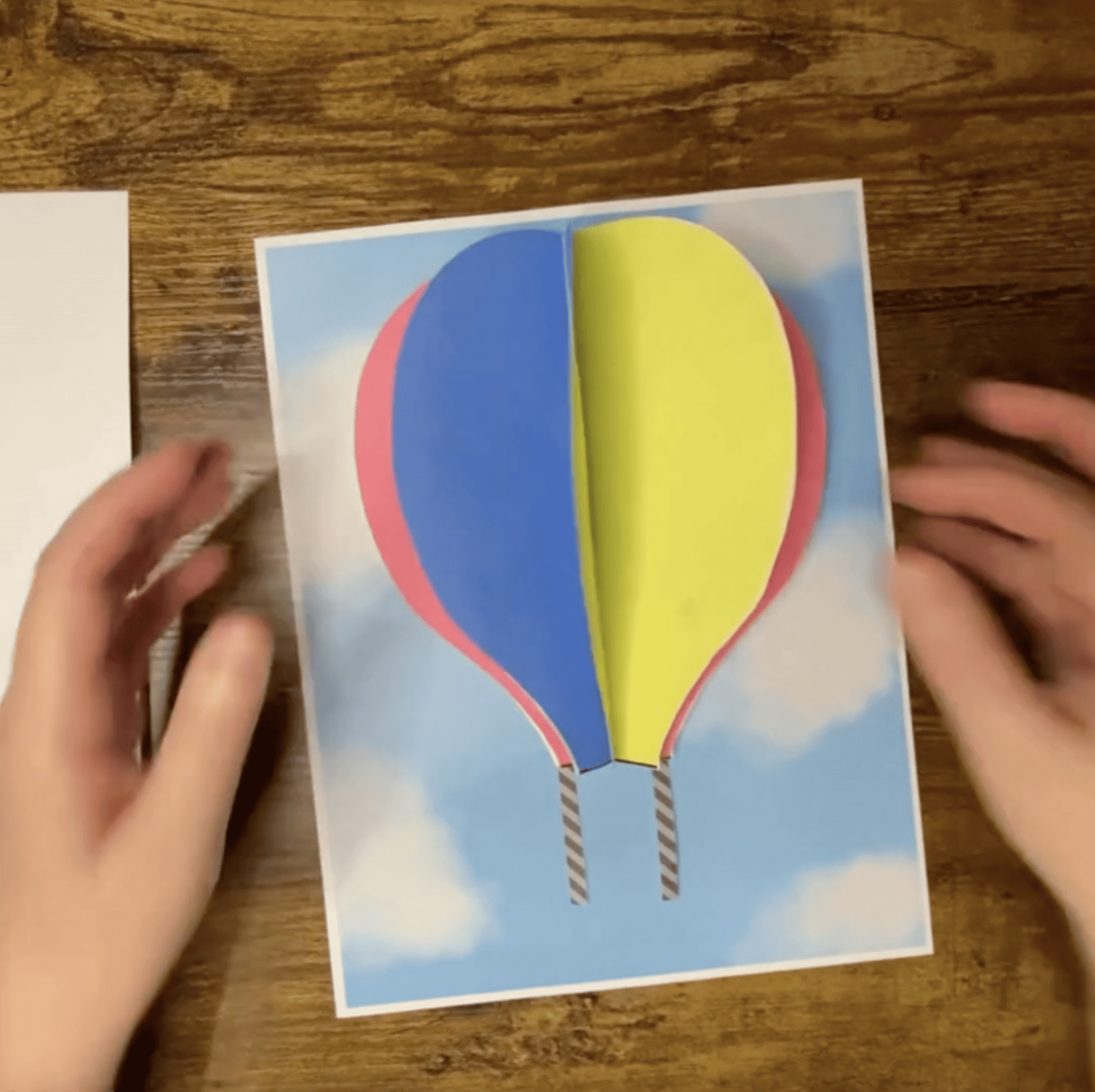 A child is working on a hot air balloon craft project, using paper to assemble a colorful balloon against a sky background, with a glue stick visible in hand.
