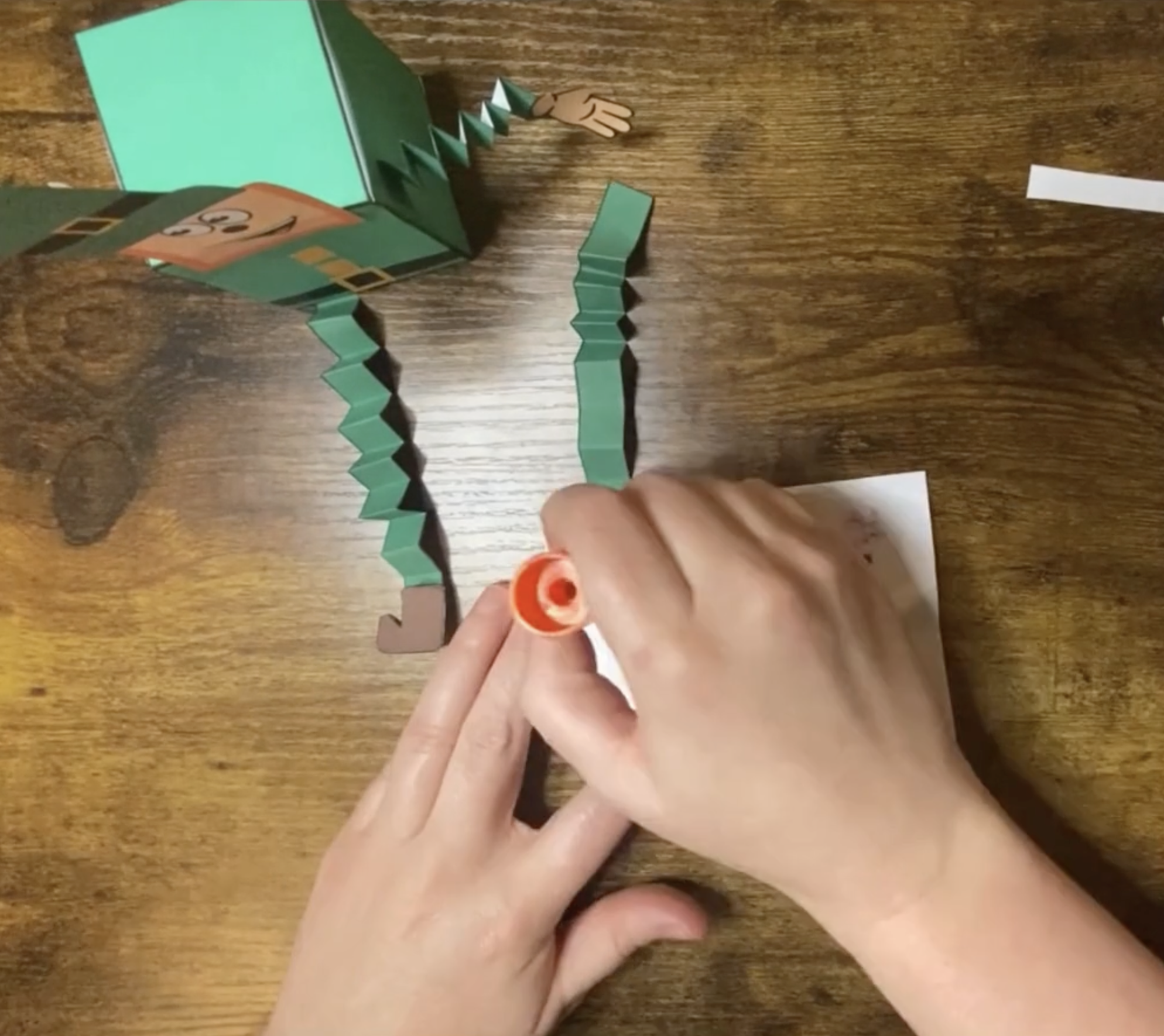 A person is assembling a green leprechaun craft from paper materials, suitable for St. Patrick’s Day activities for preschool and kindergarten children.
