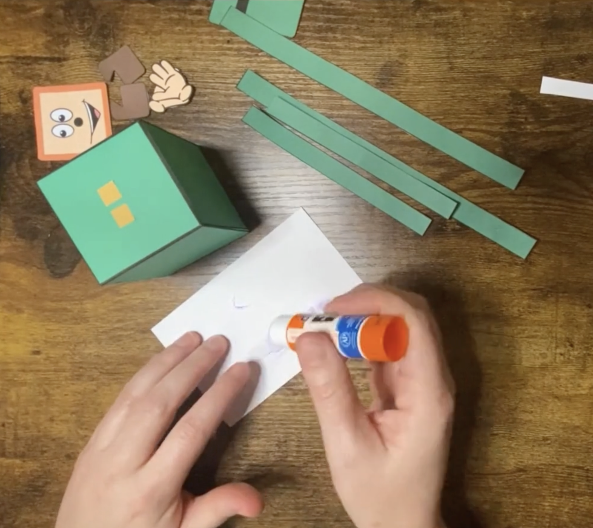 A person is applying glue to a piece of paper while surrounded by strips of green paper and a green box, which can be used for St. Patrick’s Day crafts, including an easy leprechaun craft for preschool children.
