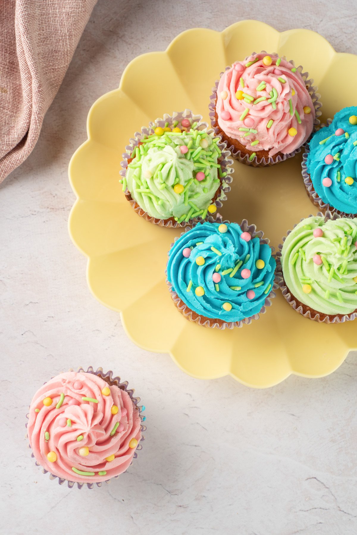 A yellow plate holds six colorful frosted cupcakes in pastel shades, with pink, green, and blue buttercream designs, decorated with sprinkles, suggesting a festive treat for Easter.
