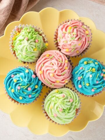 A plate of six colorful cupcakes, decorated with pastel buttercream frosting in pink, blue, and green, topped with sprinkles, showcasing ideas for Easter cupcakes and easy gluten-free desserts.