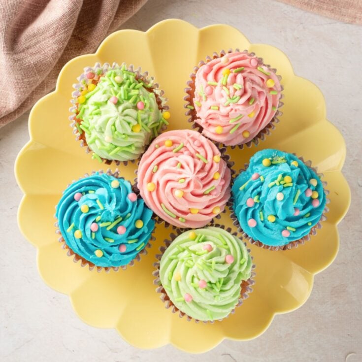 A plate of six colorful cupcakes, decorated with pastel buttercream frosting in pink, blue, and green, topped with sprinkles, showcasing ideas for Easter cupcakes and easy gluten-free desserts.