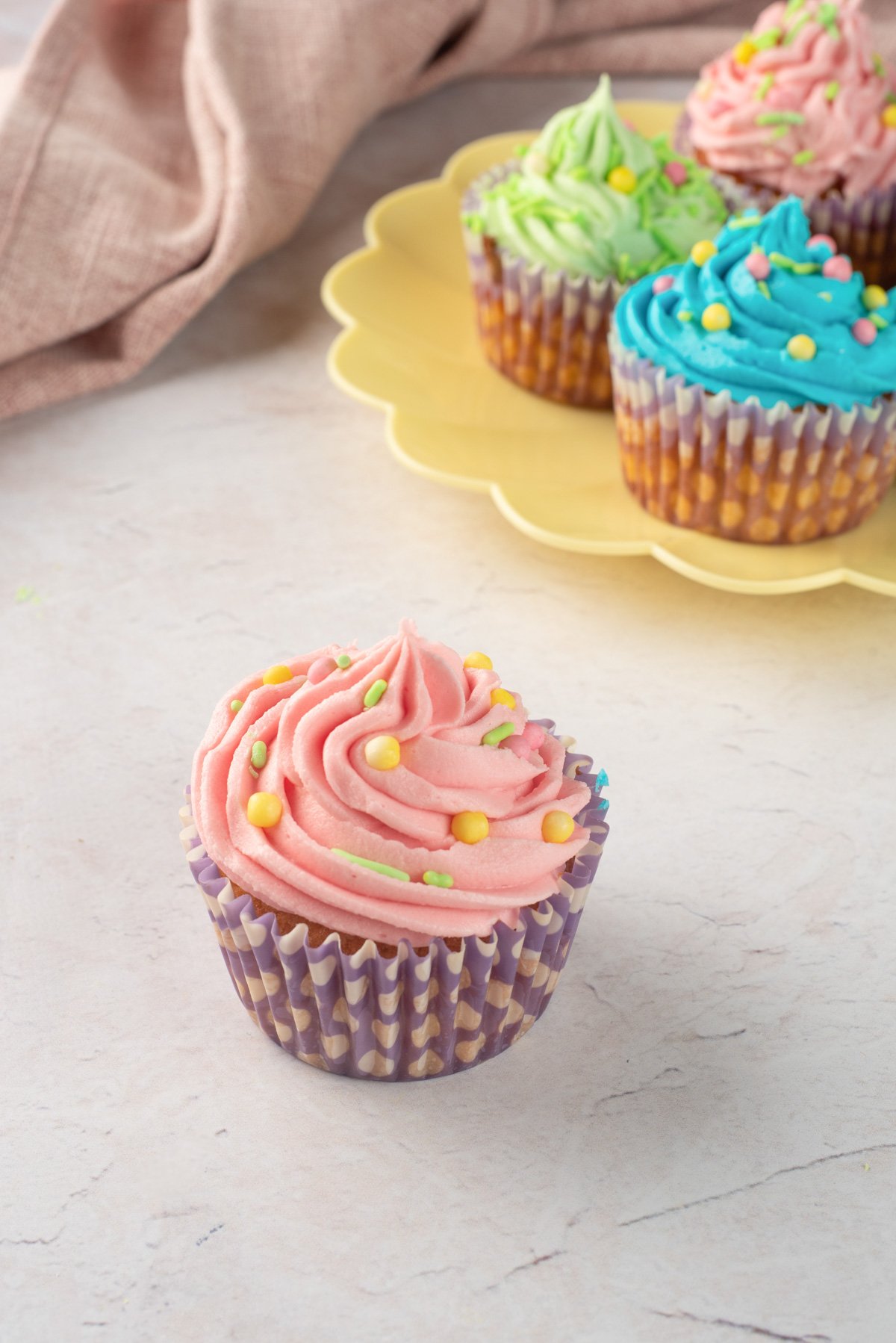 A yellow plate holds six colorful frosted cupcakes in pastel shades, with pink, green, and blue buttercream designs, decorated with sprinkles, suggesting a festive treat for Easter.
