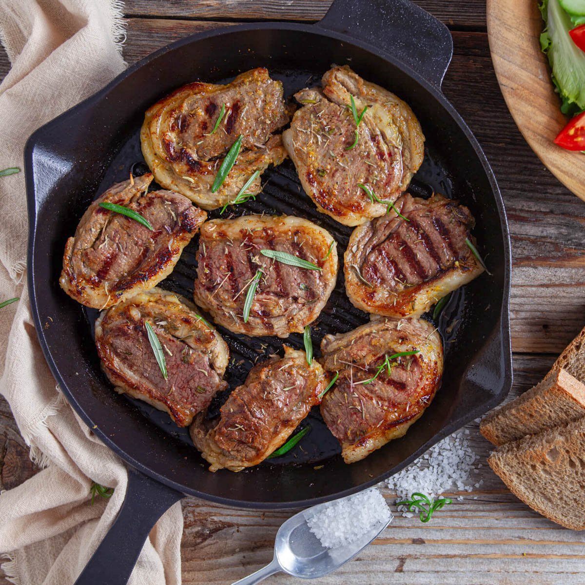 A cast iron skillet holds several herb-crusted lamb chops, seasoned and broiled