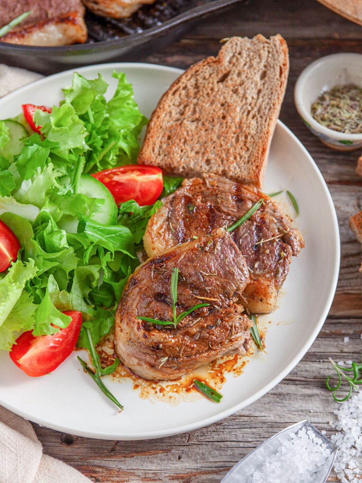 A plate of oven-roasted lamb chops garnished with herbs, served alongside a fresh salad of lettuce and tomatoes, with slices of bread in the background.
