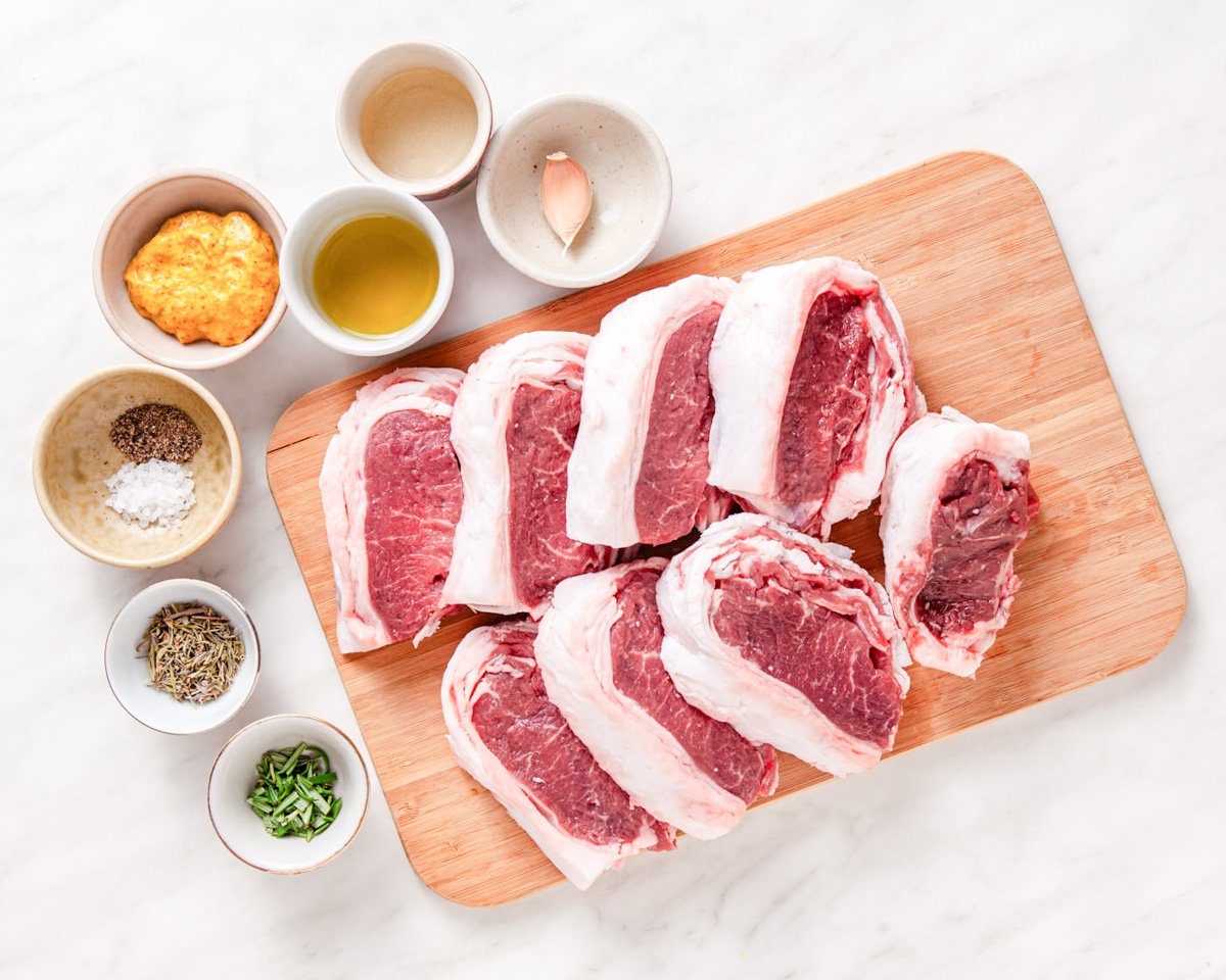 A wooden cutting board displays several uncooked lamb chops alongside small bowls of various seasonings and condiments, suggesting preparation for baked or pan-seared lamb chop recipes.
