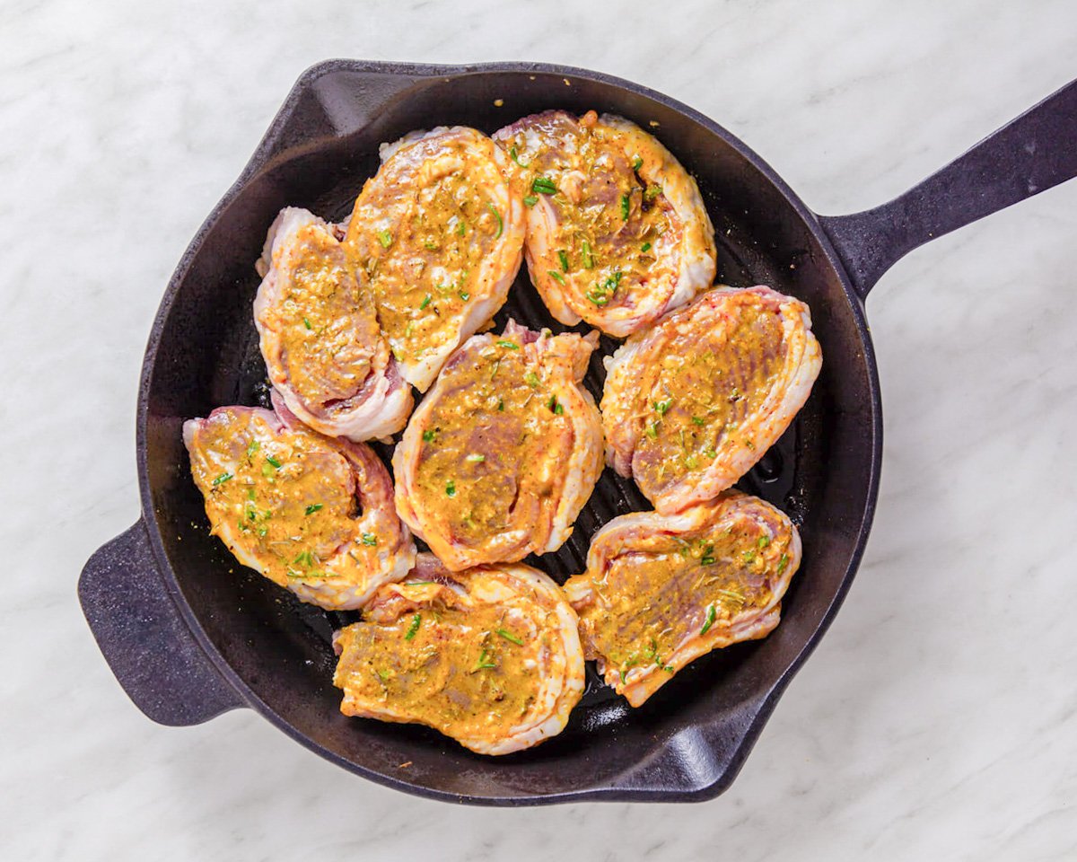 A cast iron skillet holds several herb-crusted lamb chops, seasoned and ready for cooking.
