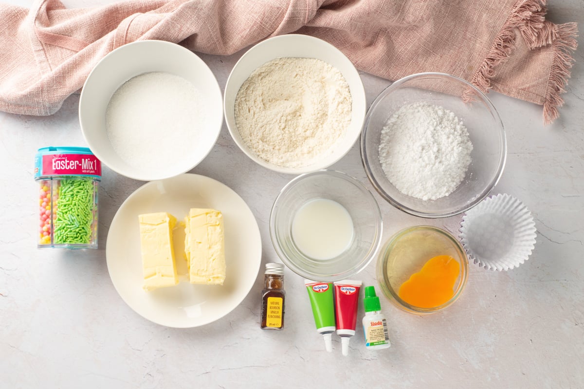 An array of baking ingredients including bowls of sugar, flour, and butter, a container of Easter-themed sprinkles, and various liquids and food colorings, set against a light background, suggesting preparations for gluten-free vanilla cupcakes for Easter.
