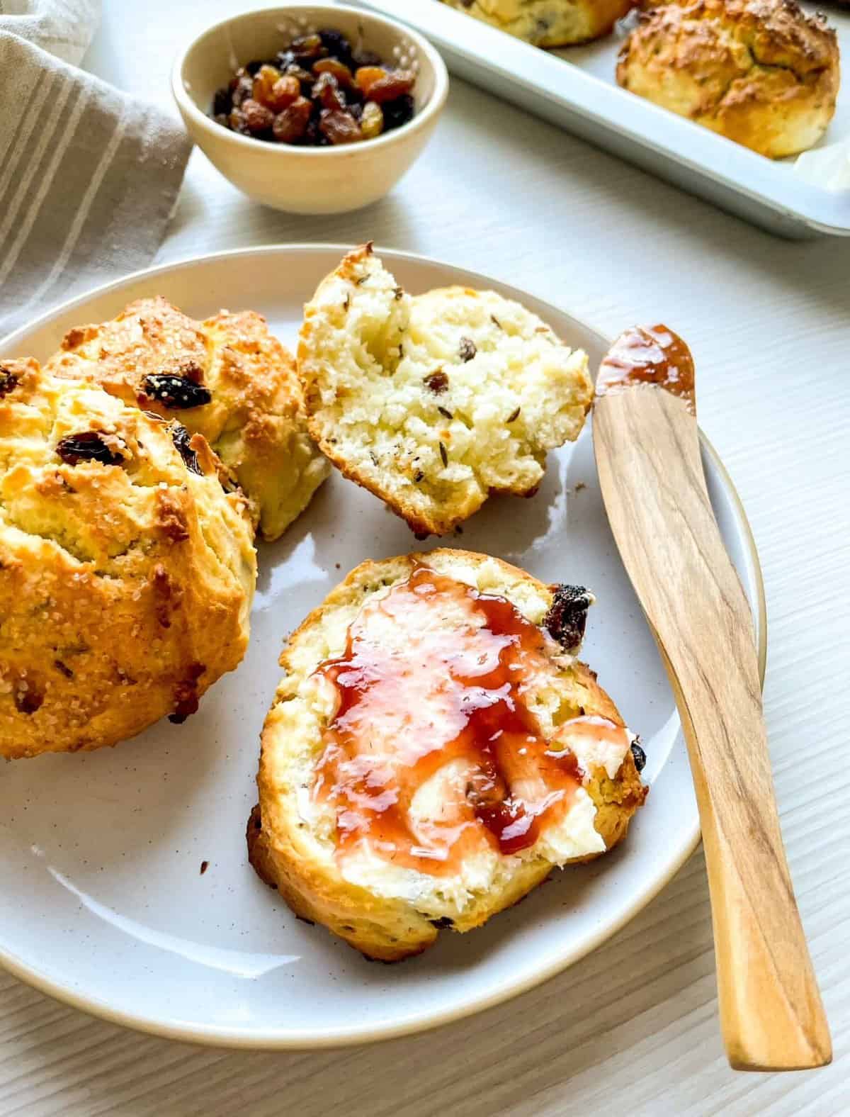 A plate of freshly baked Irish soda bread scones with raisins, featuring a portion spread with butter and topped with jam, accompanied by a small bowl of mixed dried fruits.
