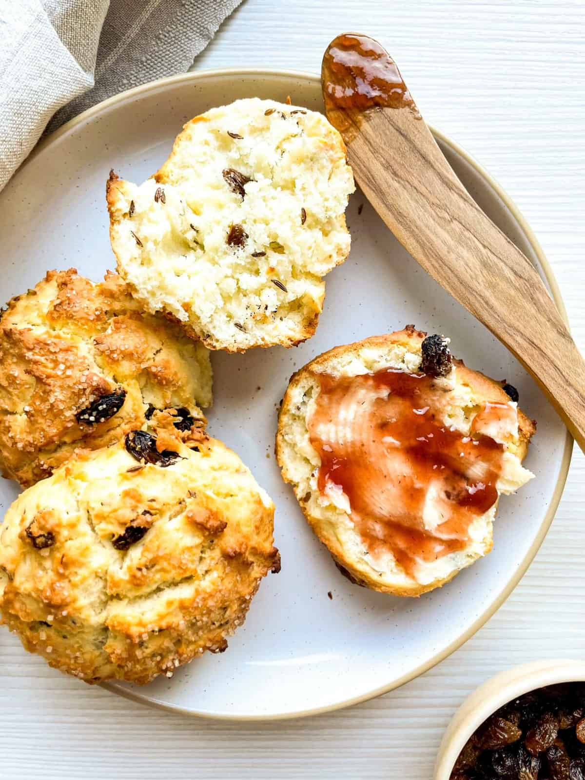 A plate of freshly baked Irish soda bread scones, some cut open to reveal a soft interior with raisins, accompanied by a wooden butter knife and a small bowl of raisins.
