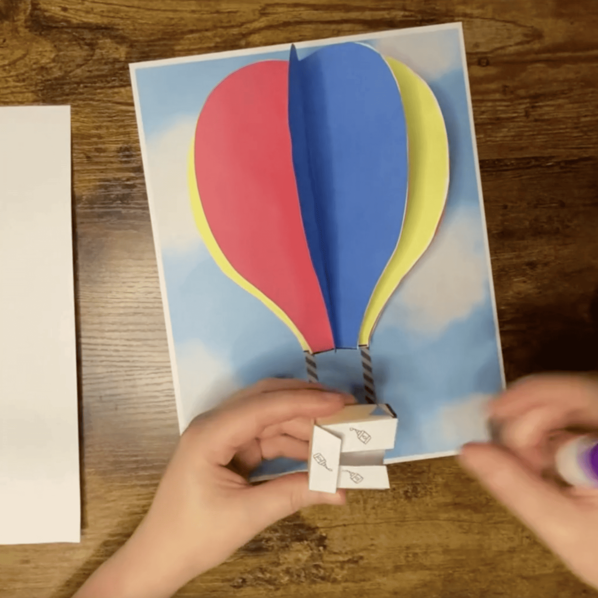 A paper hot air balloon craft with colorful balloon shapes is displayed against a sky background, illustrating a preschool art project.
