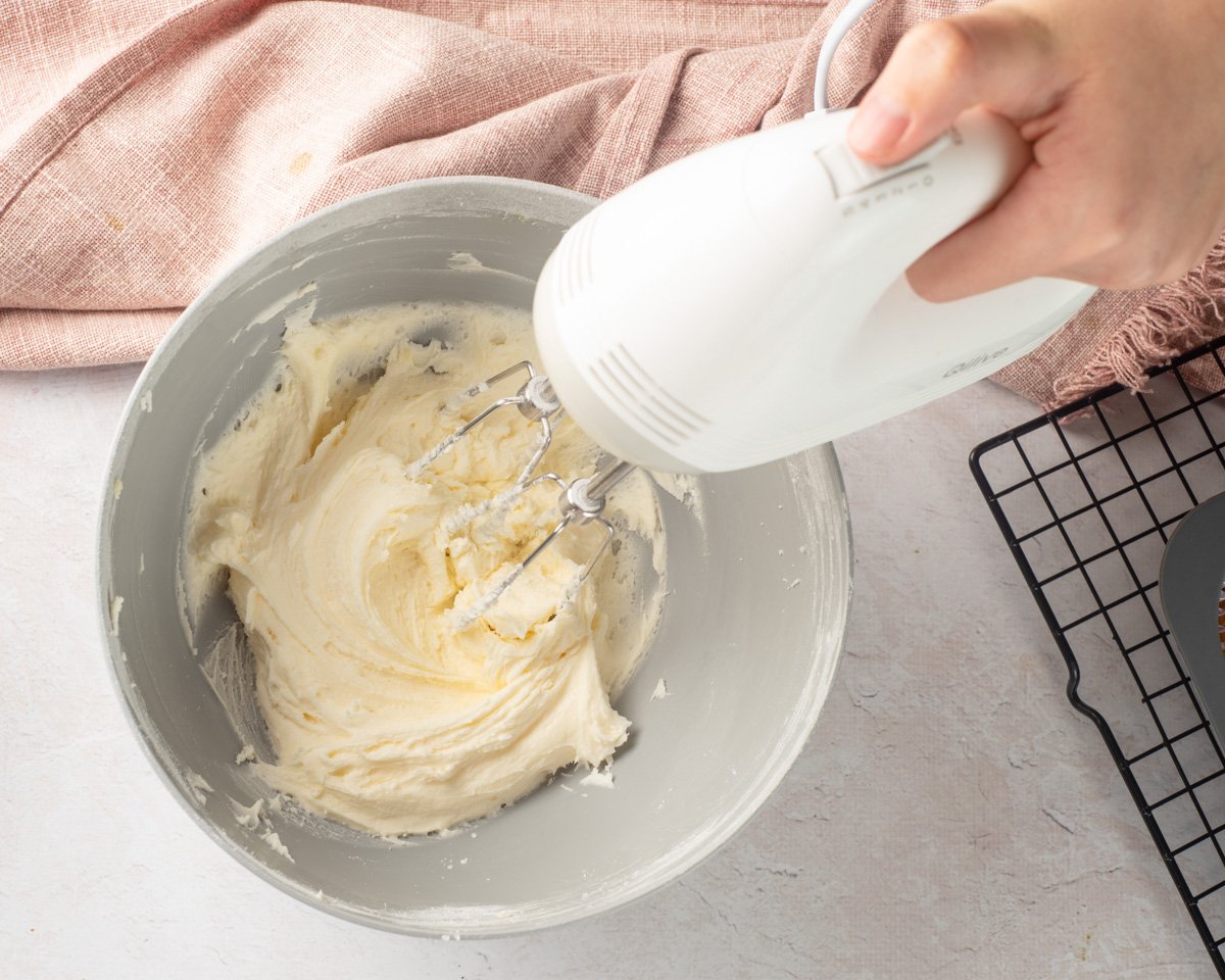 A hand using a hand mixer is blending buttercream in a gray mixing bowl, with a pink cloth and cooling rack in the background, suggesting preparation for gluten free cupcakes suitable for Easter.
