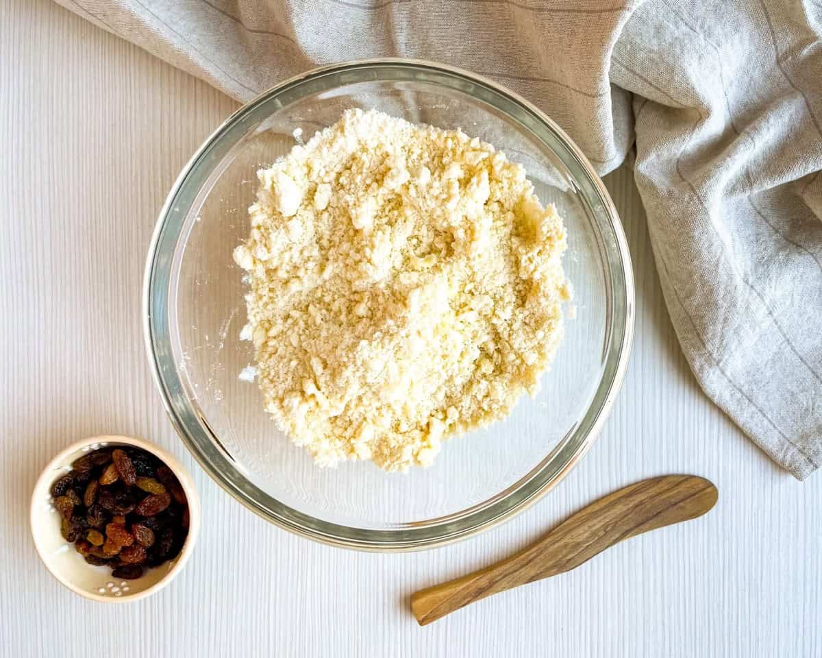 A clear glass bowl containing a crumbly mixture sits next to a small bowl of raisins and a wooden spoon on a light surface, suggesting preparation for an easy buttermilk scone or Irish soda bread recipe.
