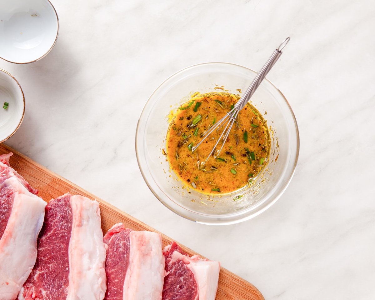 A wooden cutting board with raw lamb chops beside a bowl of mixed herb marinade on a marble surface.
