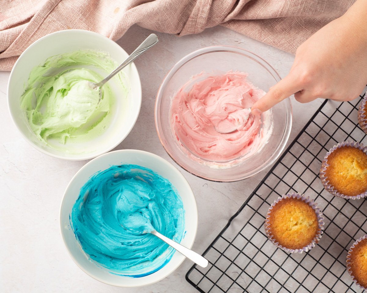 A hand is seen mixing pink buttercream icing in a bowl, surrounded by bowls of green and blue frosting, with cooling gluten-free vanilla cupcakes on a wire rack nearby.
