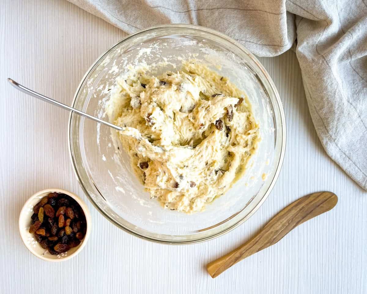 A glass bowl contains a sticky dough mixed with raisins, accompanied by a small bowl of mixed dried fruits and a butter knife on a light surface, suggesting preparation for buttermilk scones or Irish soda bread.
